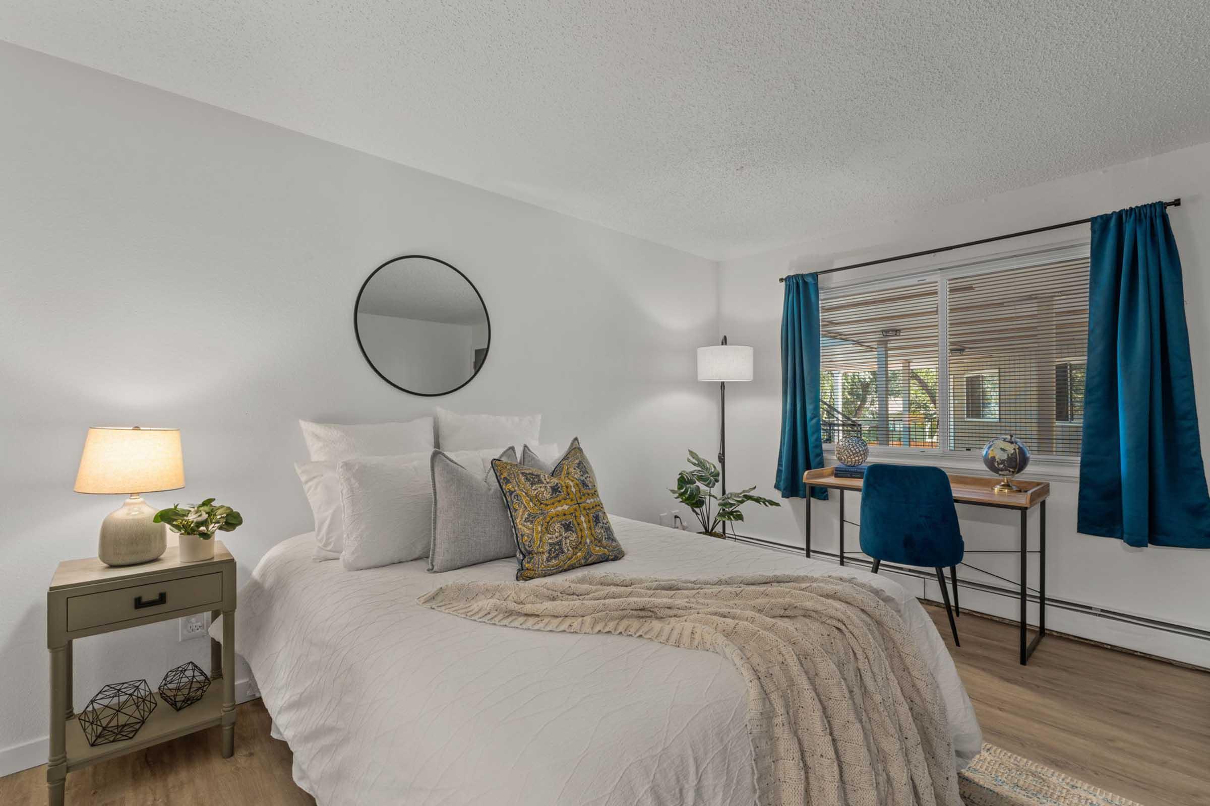 A modern bedroom featuring a queen-sized bed with white linens and decorative pillows. There's a nightstand with a lamp, a round mirror on the wall, and a workspace with a desk and blue chair. Large window with blue curtains lets in natural light, creating a bright and inviting atmosphere.