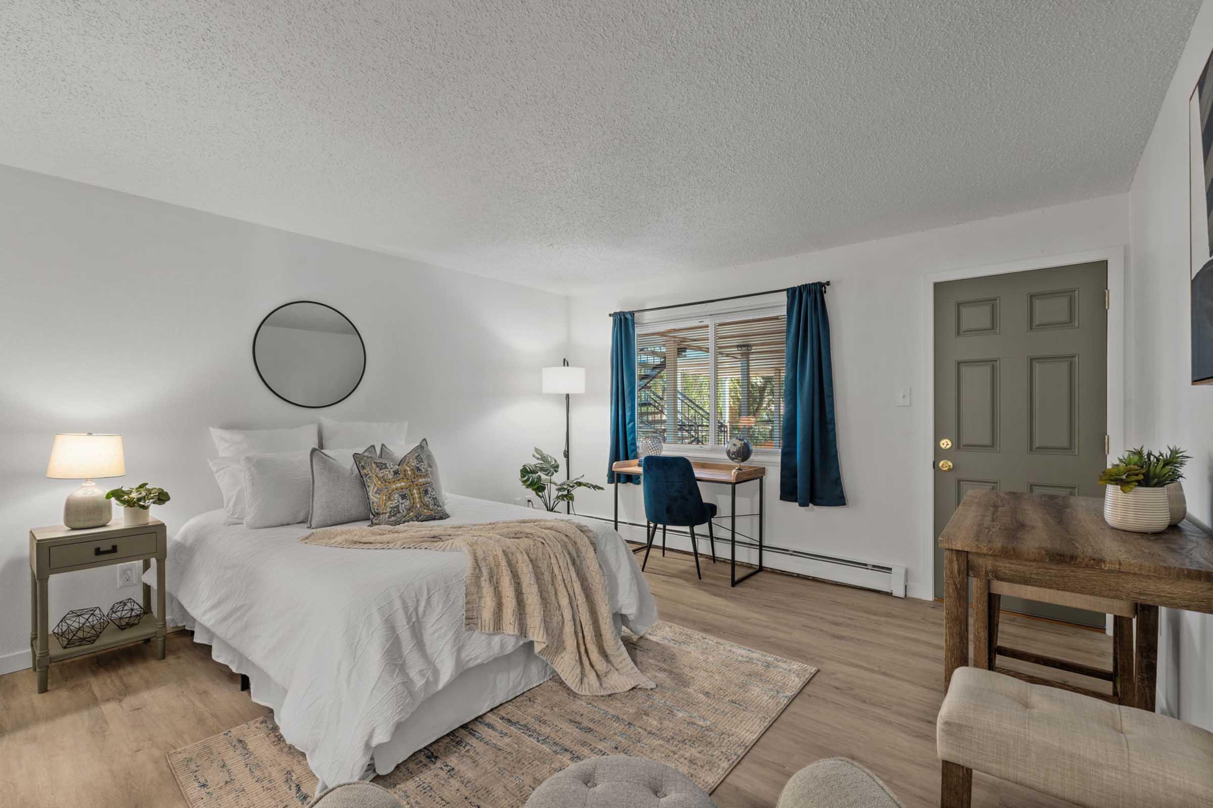 A cozy bedroom featuring a queen-sized bed with white linens and decorative pillows. There's a small side table with a lamp, a window with blue curtains letting in natural light, and a workspace with a chair and desk. A plant and soft decor elements enhance the inviting atmosphere.