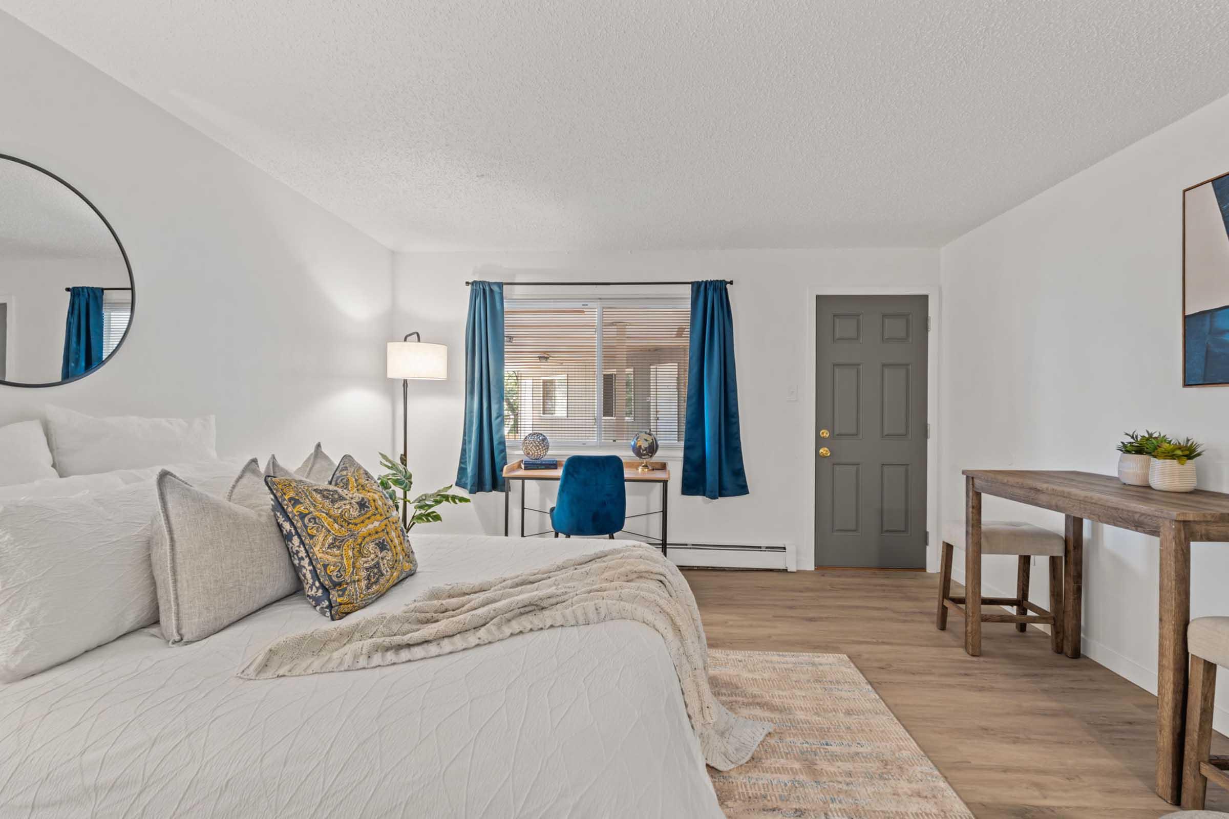 Bright and airy bedroom featuring a large bed with decorative pillows, a round mirror on the wall, and two blue curtains framing a window. A wooden table with a blue chair is positioned near the window, and a plant is placed on the table. The room has a neutral color scheme and warm wood flooring.