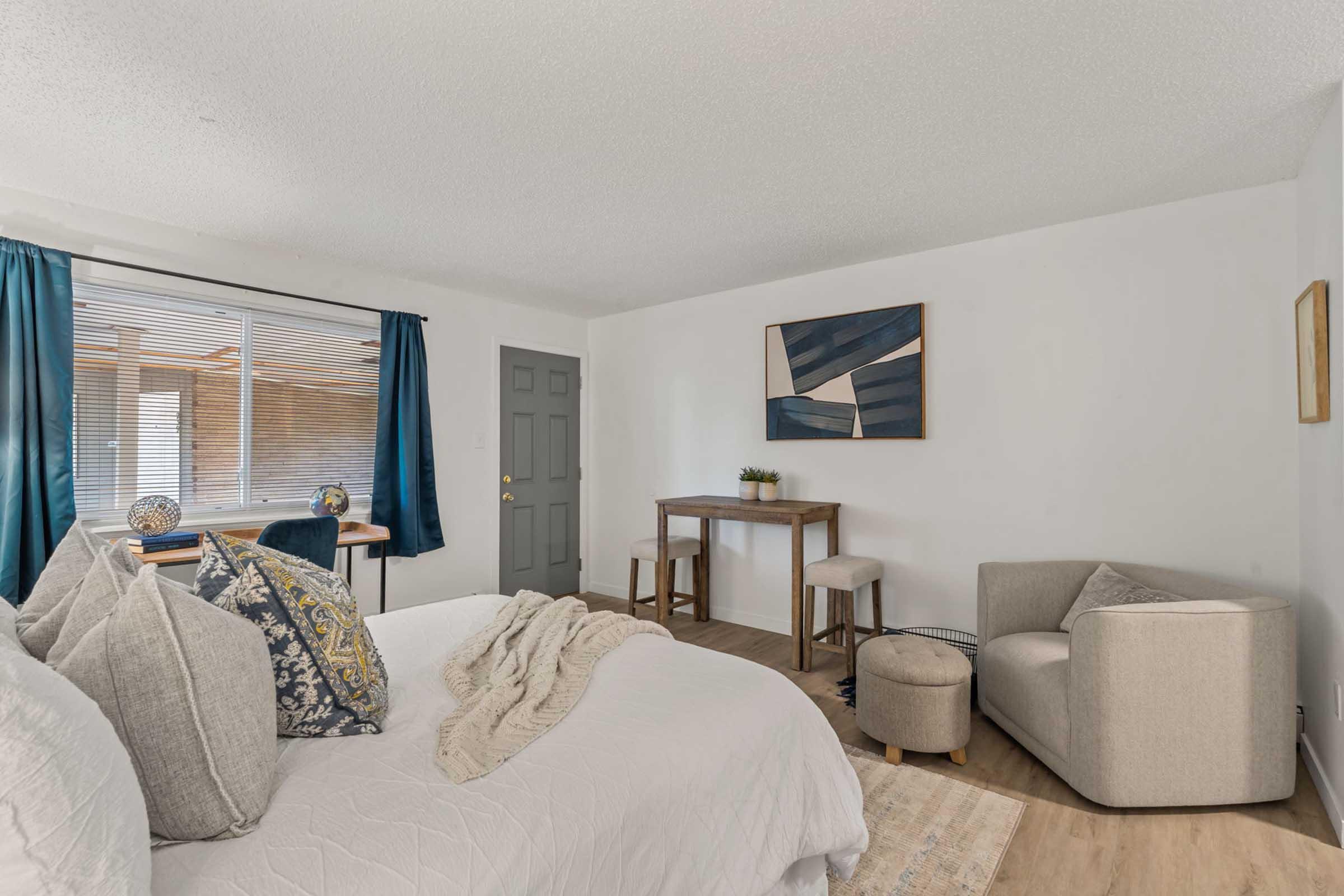 Cozy living space featuring a white bed with patterned pillows, a small wooden dining table, and a light greige armchair. Natural light streams through a window with blue curtains, complemented by a modern wall art piece. The floor is wooden, adding warmth to the minimalist decor.