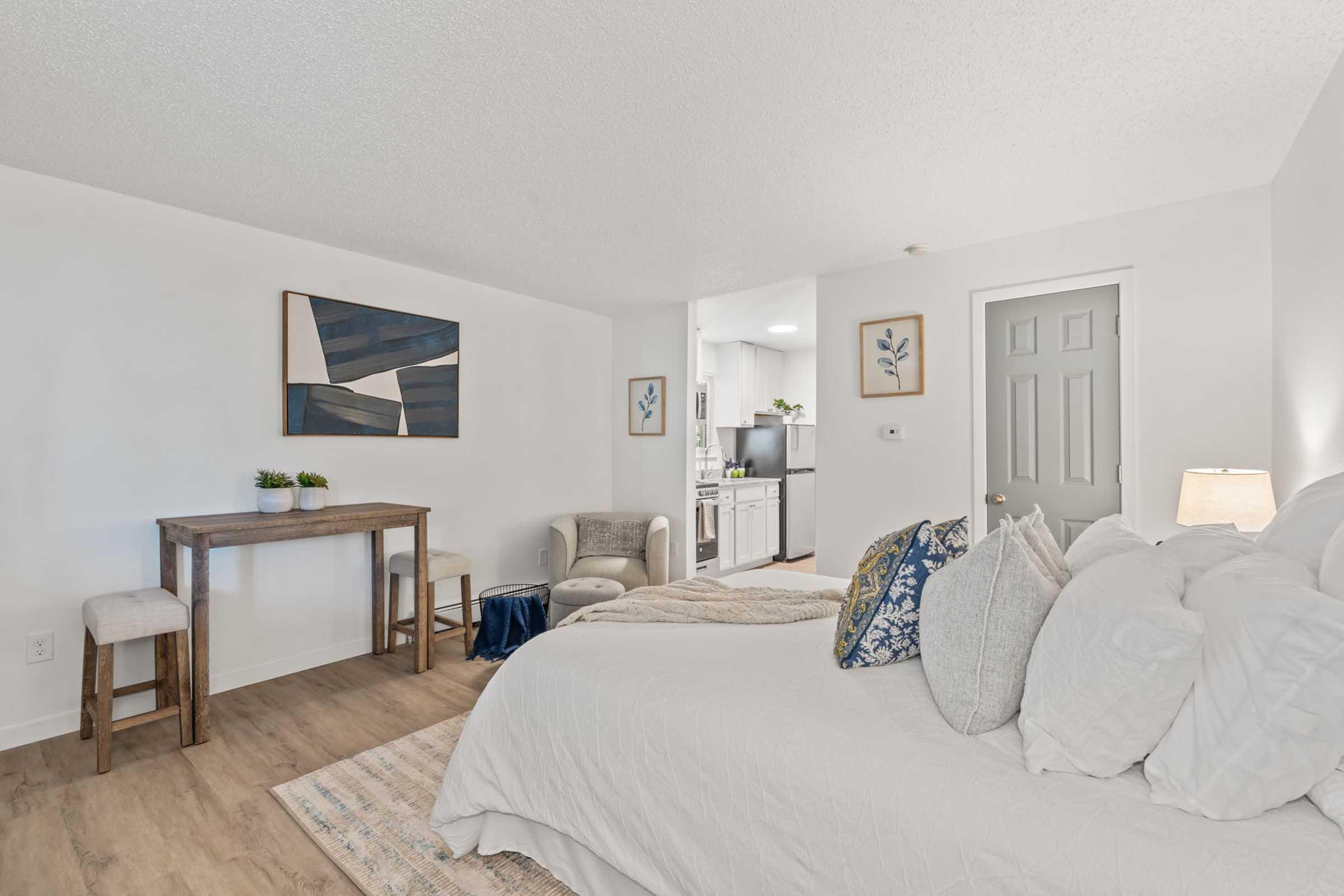 Cozy bedroom featuring a large bed with decorative pillows, a wooden table with two stools, and a comfortable chair. The room has light-colored walls, a piece of abstract art on the wall, and a small plant. A kitchen area is visible in the background, adding to the functional design.