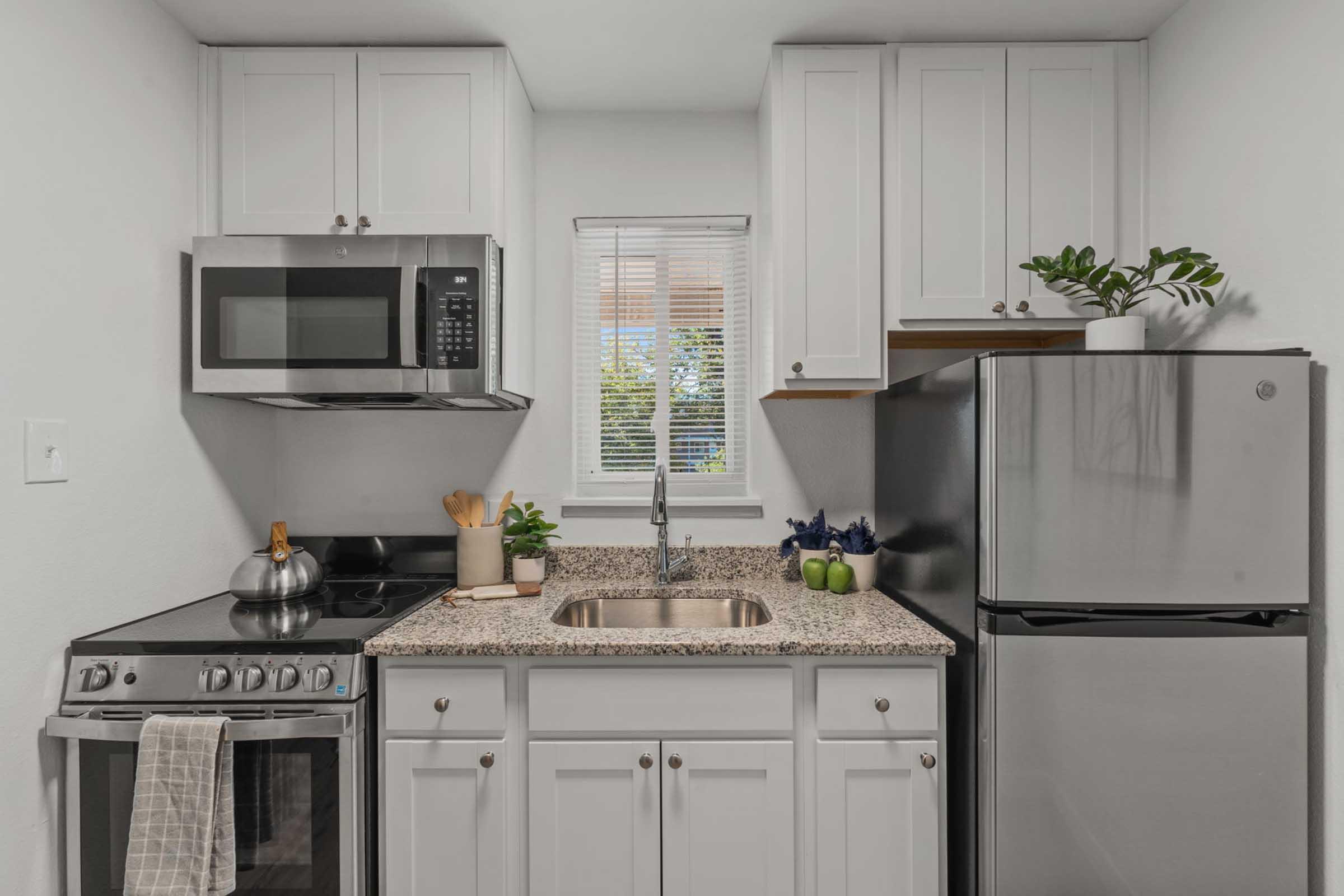 A compact kitchen featuring white cabinets, a stainless steel refrigerator, a modern stove, and a microwave. The countertop is made of granite, with kitchen utensils and a potted plant displayed. A window provides natural light, enhancing the cozy atmosphere.