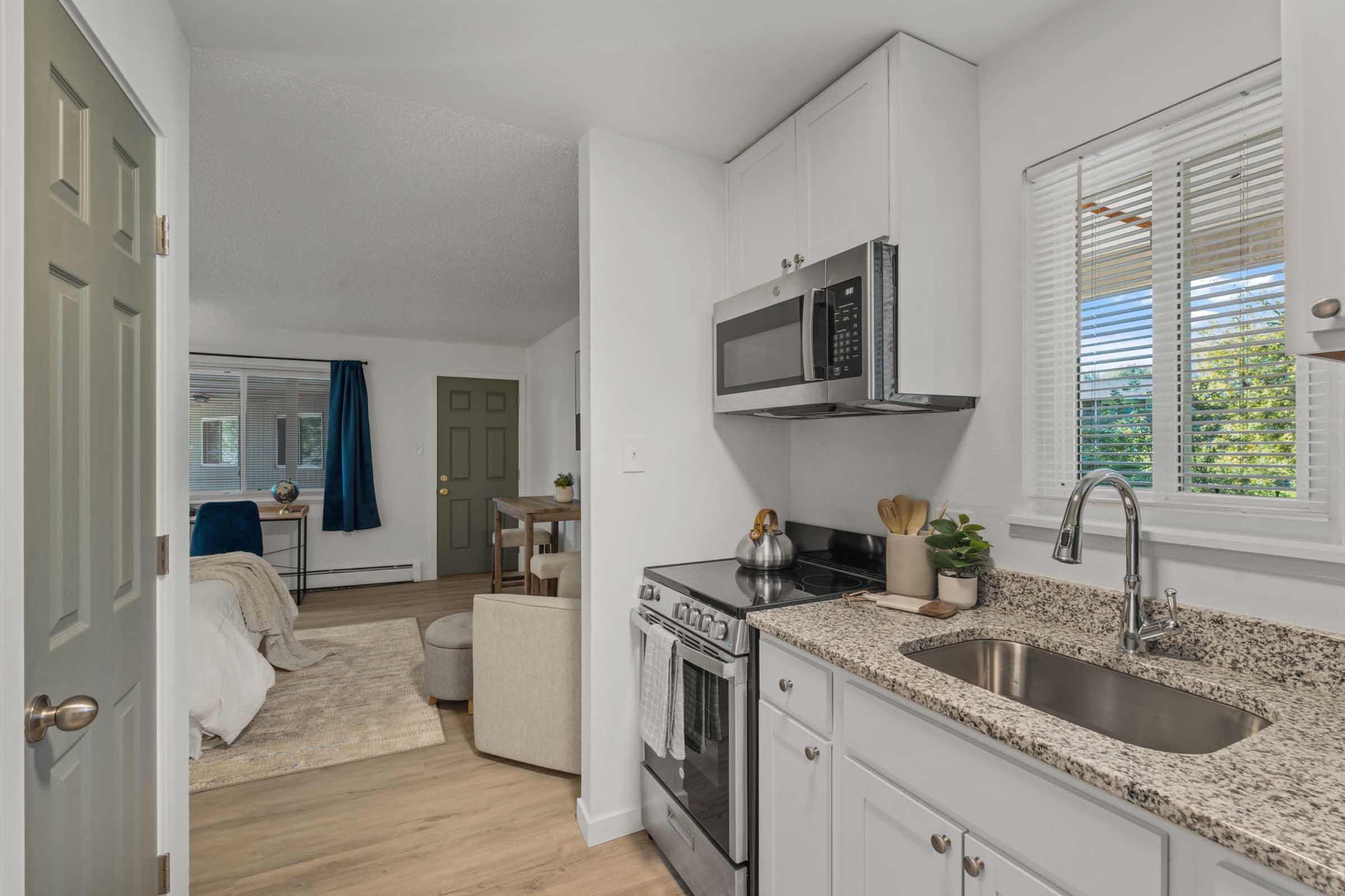 A modern kitchen featuring granite countertops, a stainless steel microwave and oven, a sink with a modern faucet, and a window allowing natural light. Adjacent to the kitchen, a living area with a desk, chair, and a cozy, unmade bed can be seen, along with a closed green door.