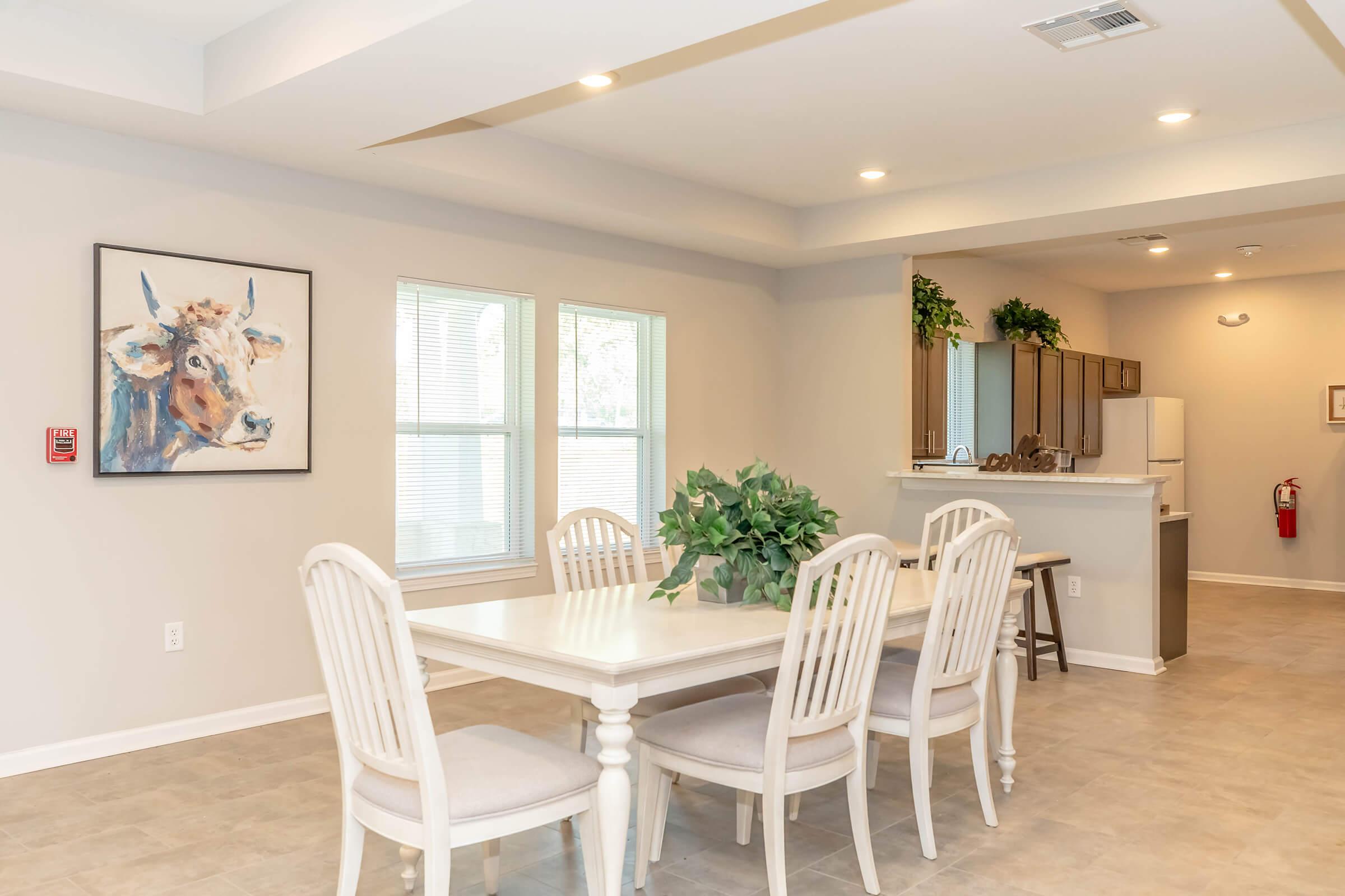 A light and airy dining area featuring a white table with six matching chairs. There is a decorative plant in the center of the table. A framed painting of a cow hangs on the wall, and the room has large windows allowing natural light to enter. The kitchen area is visible in the background.