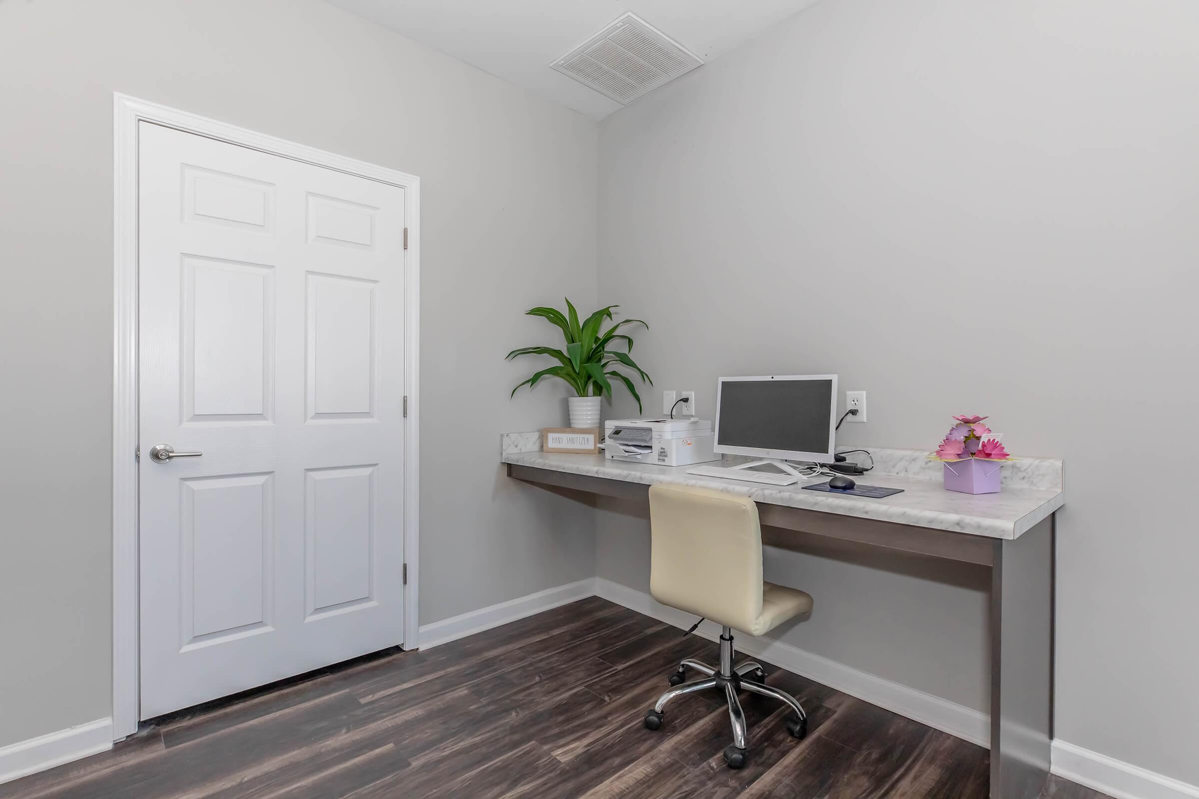 A minimalist office space featuring a small desk with a computer monitor and printer. A potted plant is placed on the desk, along with a pink flower arrangement. The walls are painted light gray, and there is a closed white door in the background. The floor is made of dark wood.