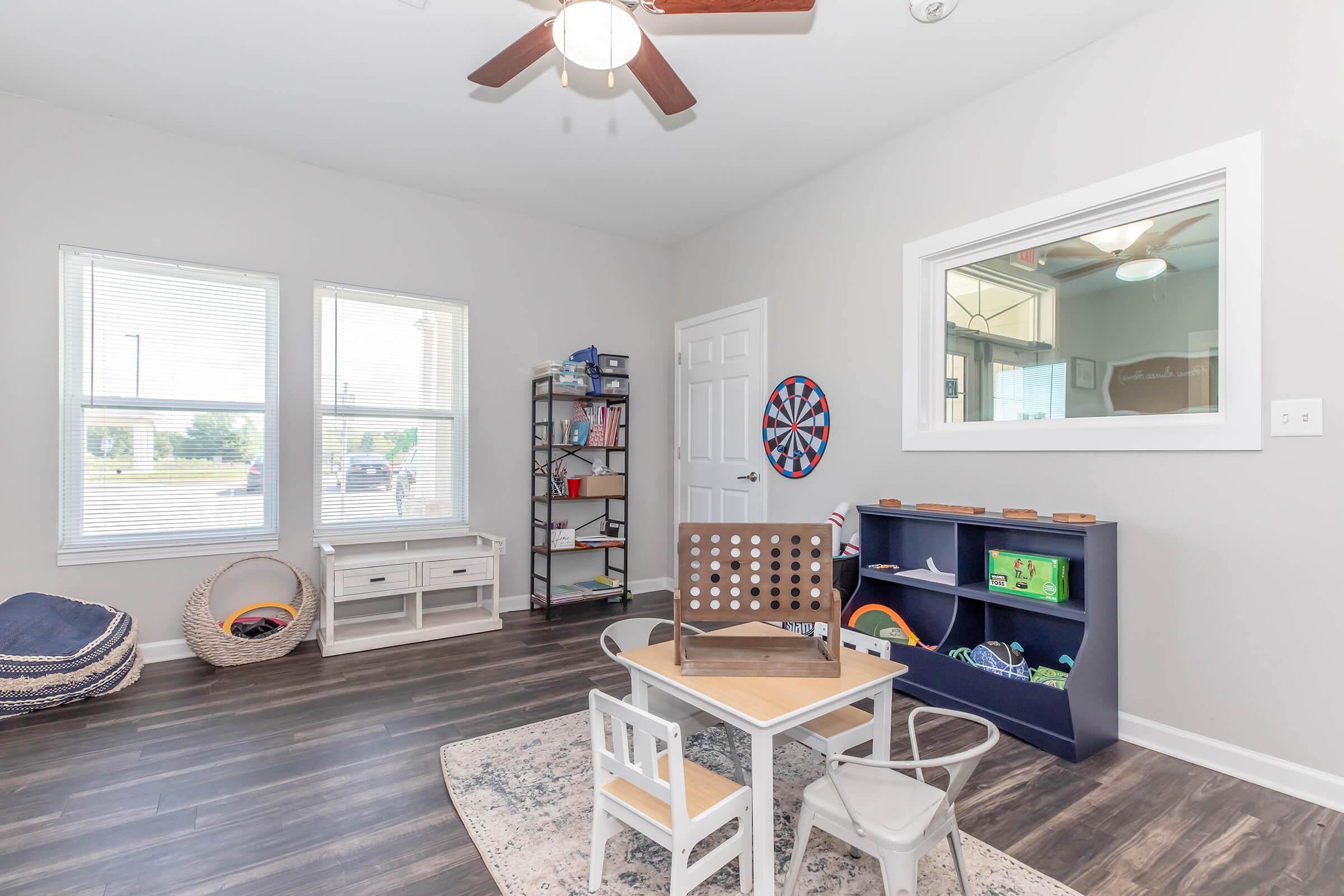 Bright playroom featuring wooden flooring, two windows, and a ceiling fan. Furnished with a small table and chairs, a blue shelf with toys, and a game board. A bookshelf is filled with books, and a dartboard is mounted on the wall. Cozy seating options include bean bags in the corner.