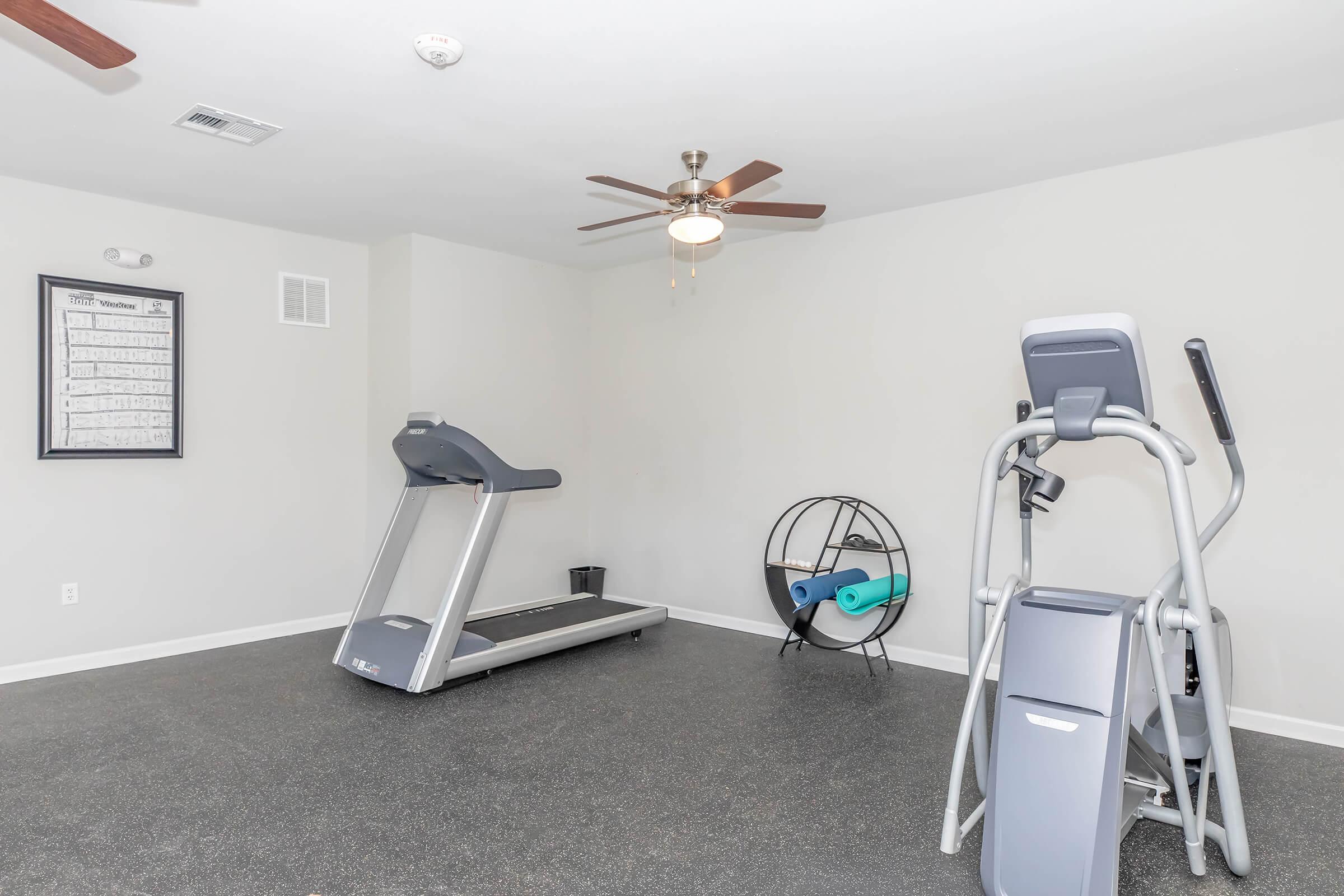 A bright, modern gym space featuring a treadmill, an elliptical machine, and a large exercise ball in a unique stand. The room has a ceiling fan and is equipped with rubber flooring. A wall-mounted calendar or schedule is visible in the background.