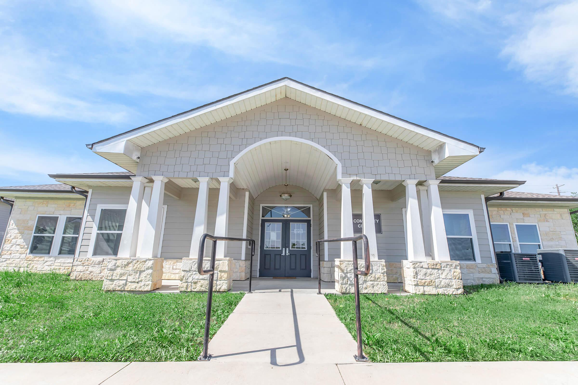A modern single-story building with a welcoming entrance, featuring a triangular roof and decorative columns. The façade has a mix of light-colored stone and siding. There are green lawns on either side of a concrete pathway leading to the front door, under a clear blue sky.