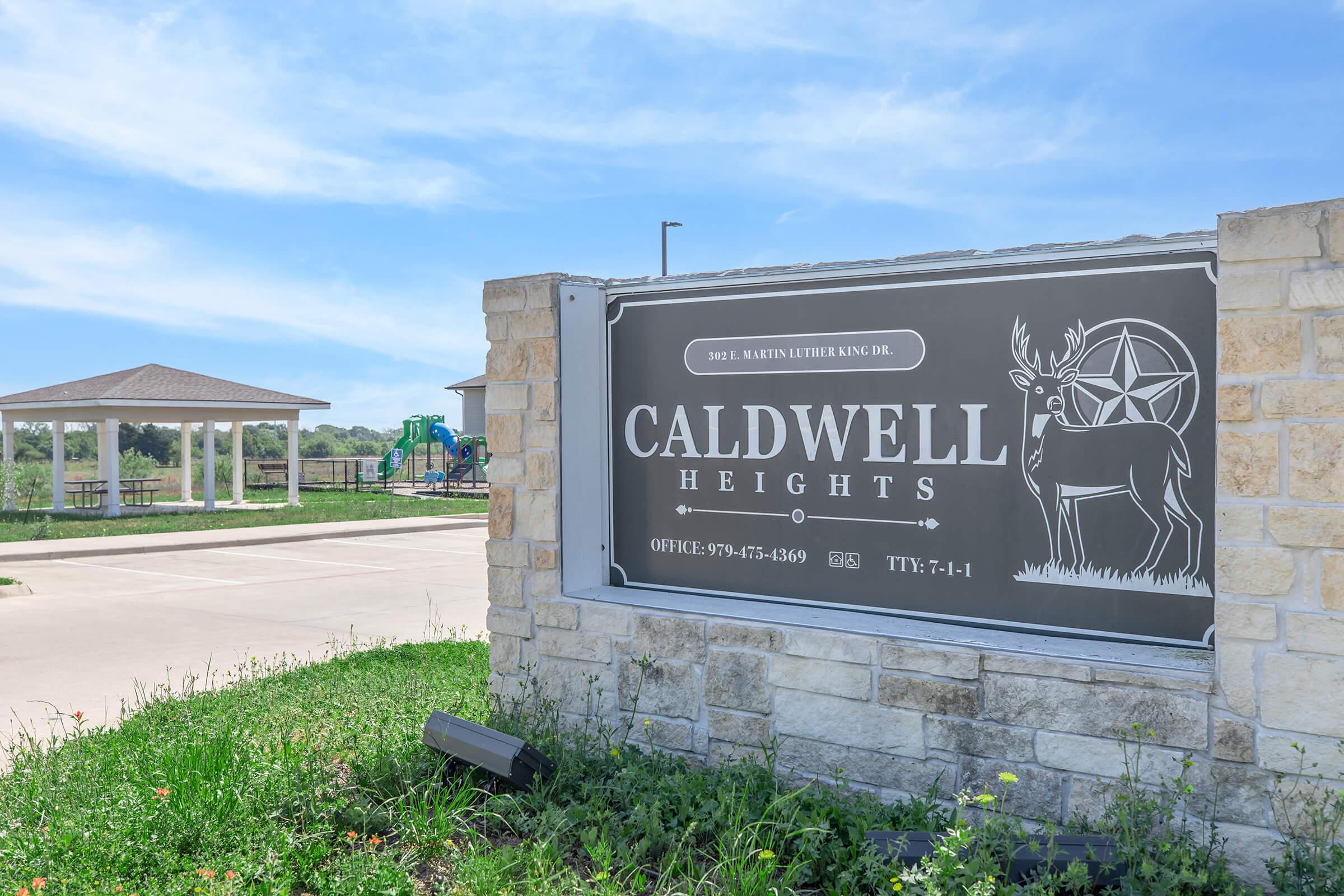 Sign for Caldwell Heights, featuring a deer and a star, surrounded by greenery. In the background, a playground and a pavilion are visible. The sign includes contact information and indicates office hours.