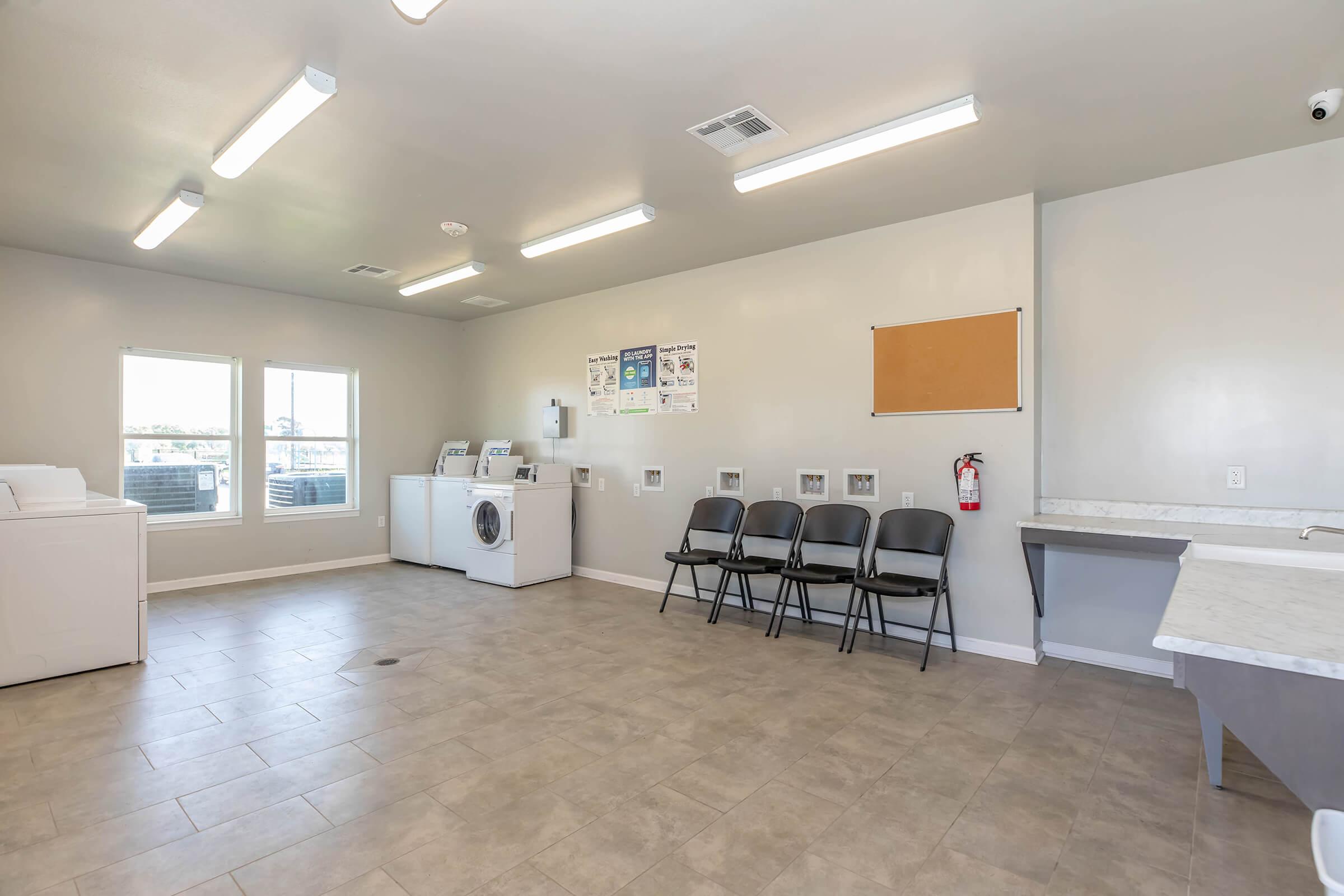 A clean laundry room featuring a washing machine, dryer, and several black chairs arranged along the wall. There are two windows letting in natural light, and the walls are painted in a neutral color. Laundry-related posters are displayed, and there's a fire extinguisher mounted nearby.