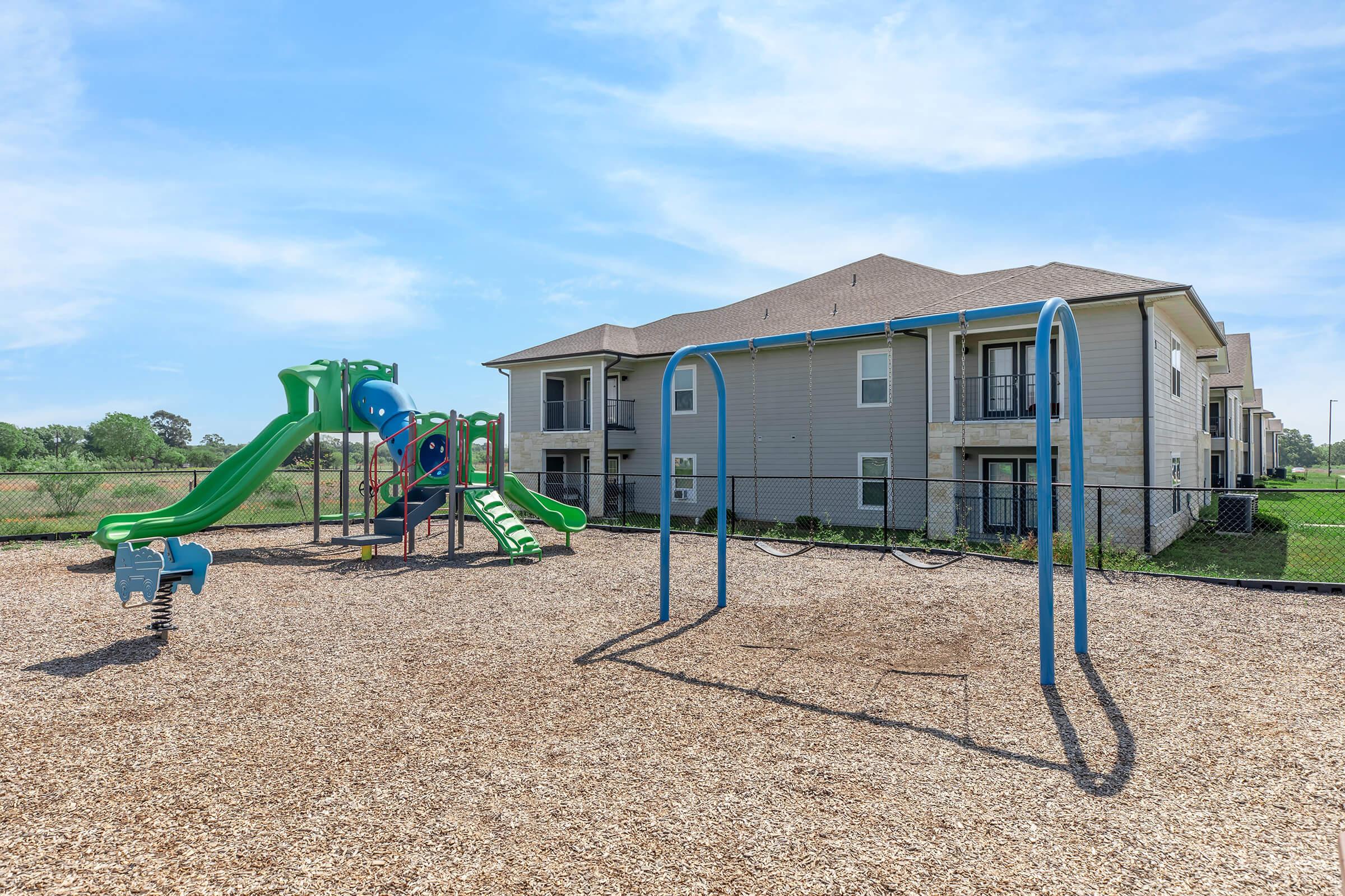 A playground featuring a green and blue slide structure, swings, and a soft wood-chip surface. In the background, an apartment building is visible, surrounded by green grass and trees under a clear blue sky.