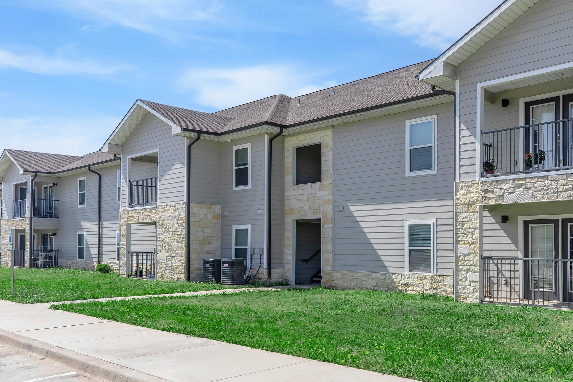 Modern two-story apartment building featuring beige siding and stone accents, with balconies on the second floor. The landscape is well-kept, with green grass and a concrete walkway leading up to the entrance. Blue sky with a few clouds overhead.
