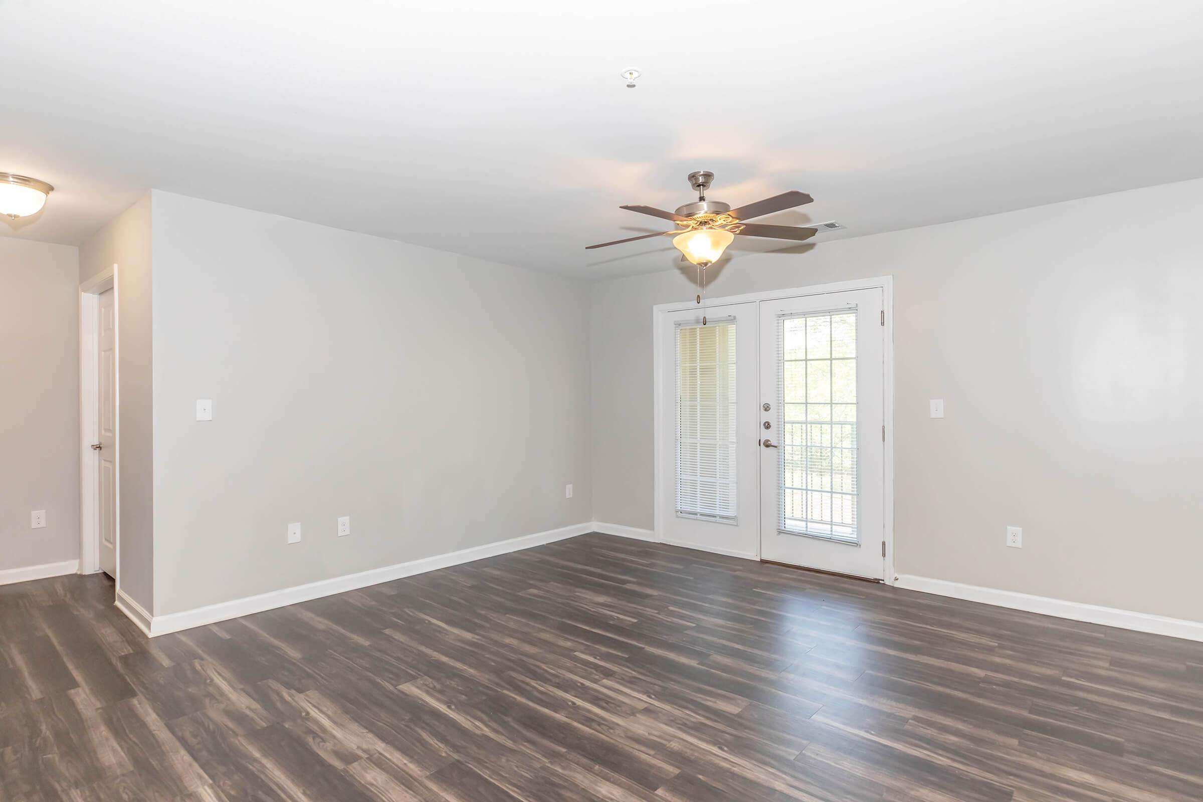 A modern, empty living room featuring light gray walls, dark hardwood floors, and a ceiling fan. There are two doorways on the left and a set of double doors leading to an outdoor area. Natural light streams through the doors, enhancing the spacious feel of the room.
