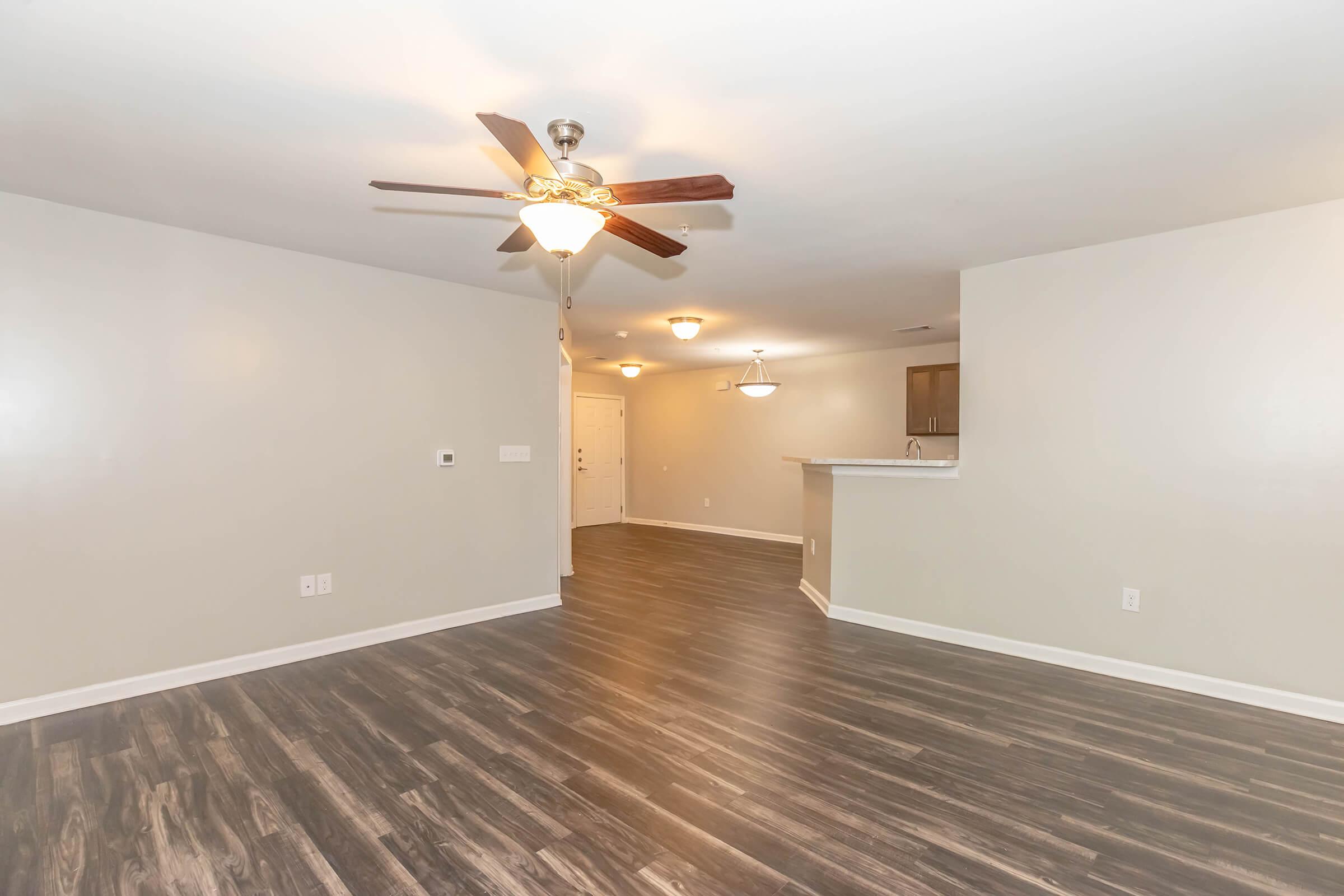 Spacious living area with a ceiling fan and light fixtures. Features a modern design with light-colored walls and dark wood-like flooring. A bar area is visible in the background, leading to a kitchen space. Natural light brightens the room, creating an inviting atmosphere.