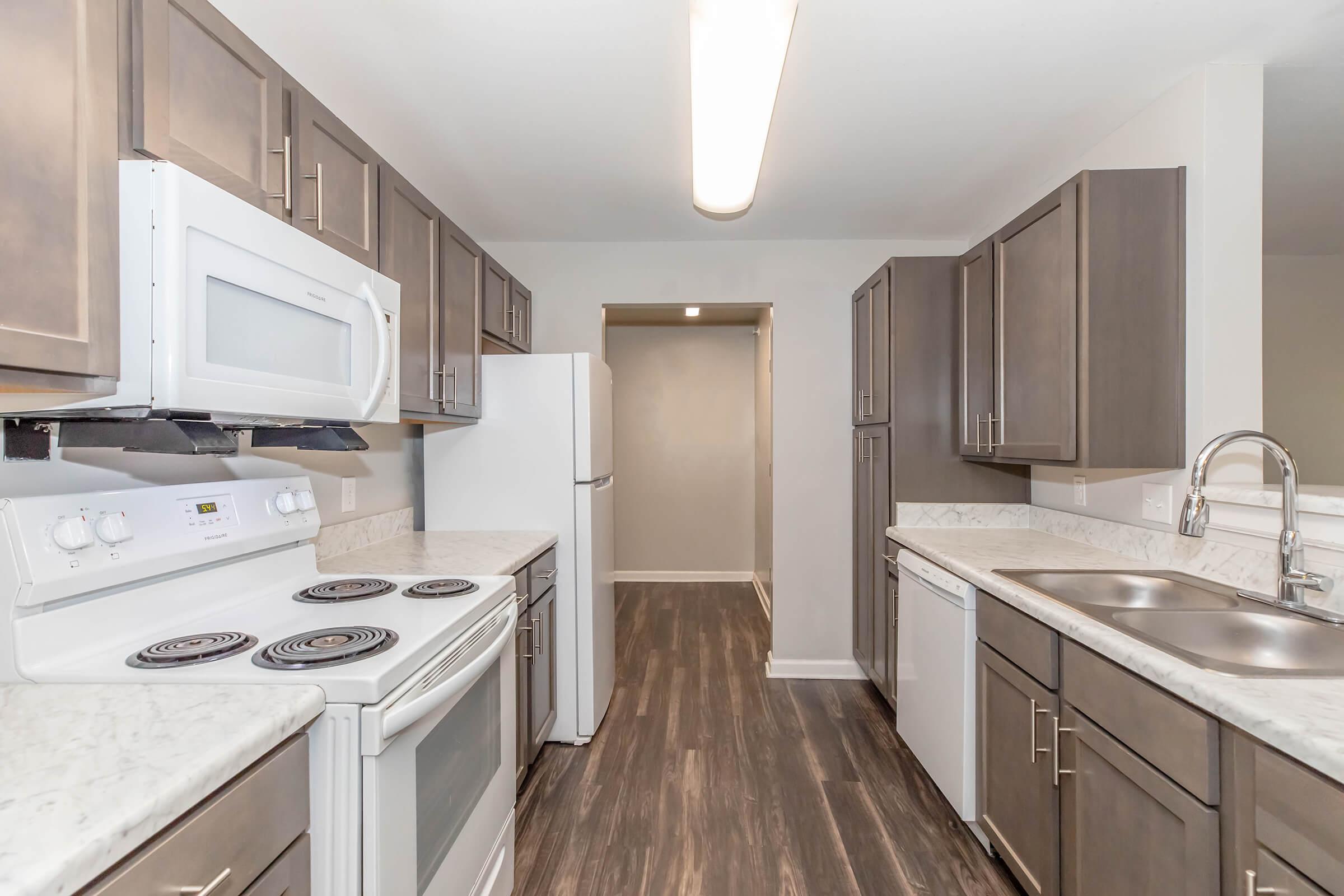 A modern kitchen featuring wooden cabinets, stainless steel appliances, a double sink, and a large countertop with a stove and microwave. The flooring is dark wood, and there is ample natural light illuminating the space. An archway leads to another room in the background.