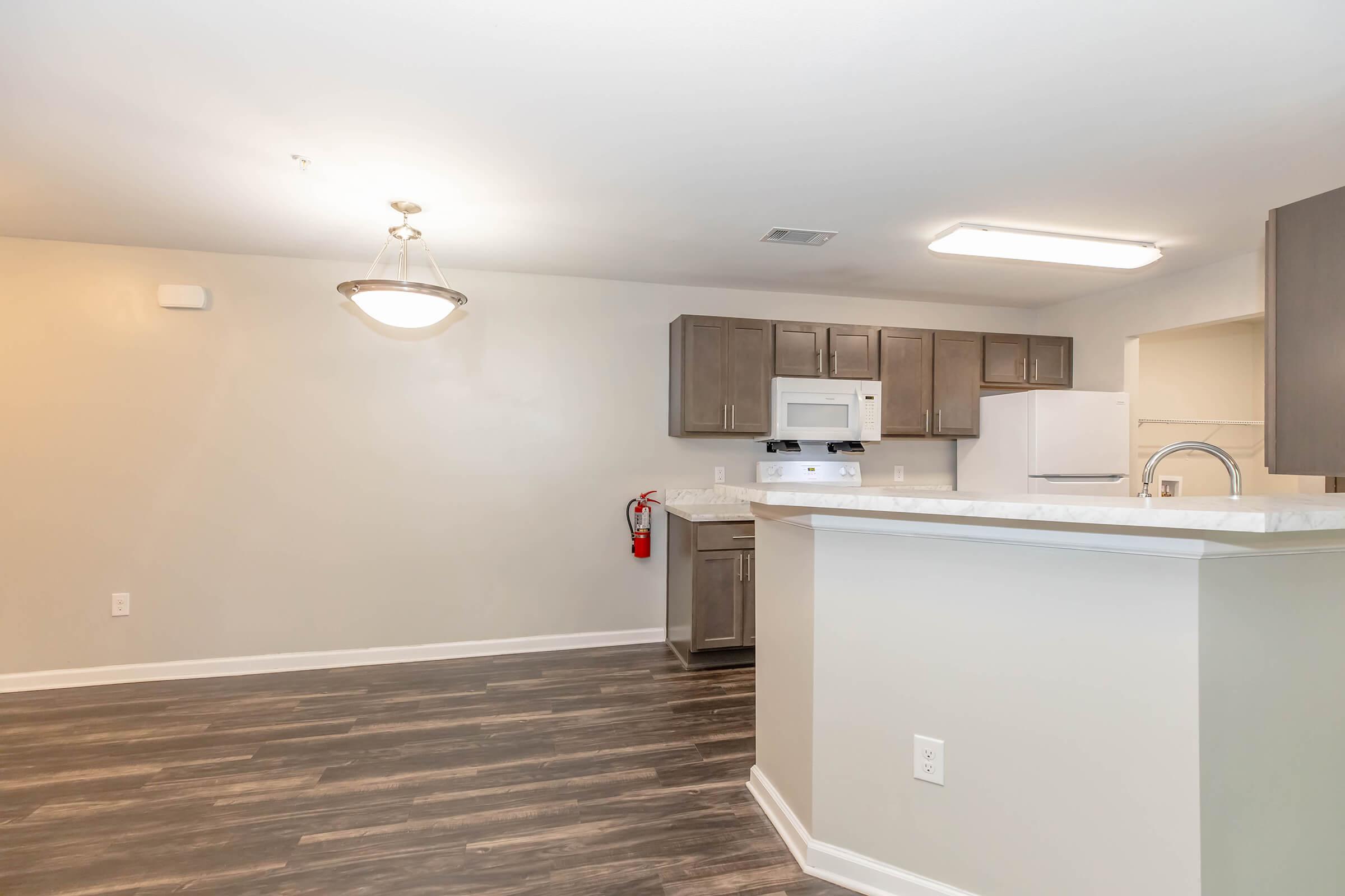 Interior view of a modern kitchen and living area featuring light-colored walls, wooden laminate flooring, and recessed lighting. The kitchen includes dark cabinets, a white microwave, and a sink with a faucet. A fire extinguisher is mounted on the wall, and there's a chandelier hanging from the ceiling.