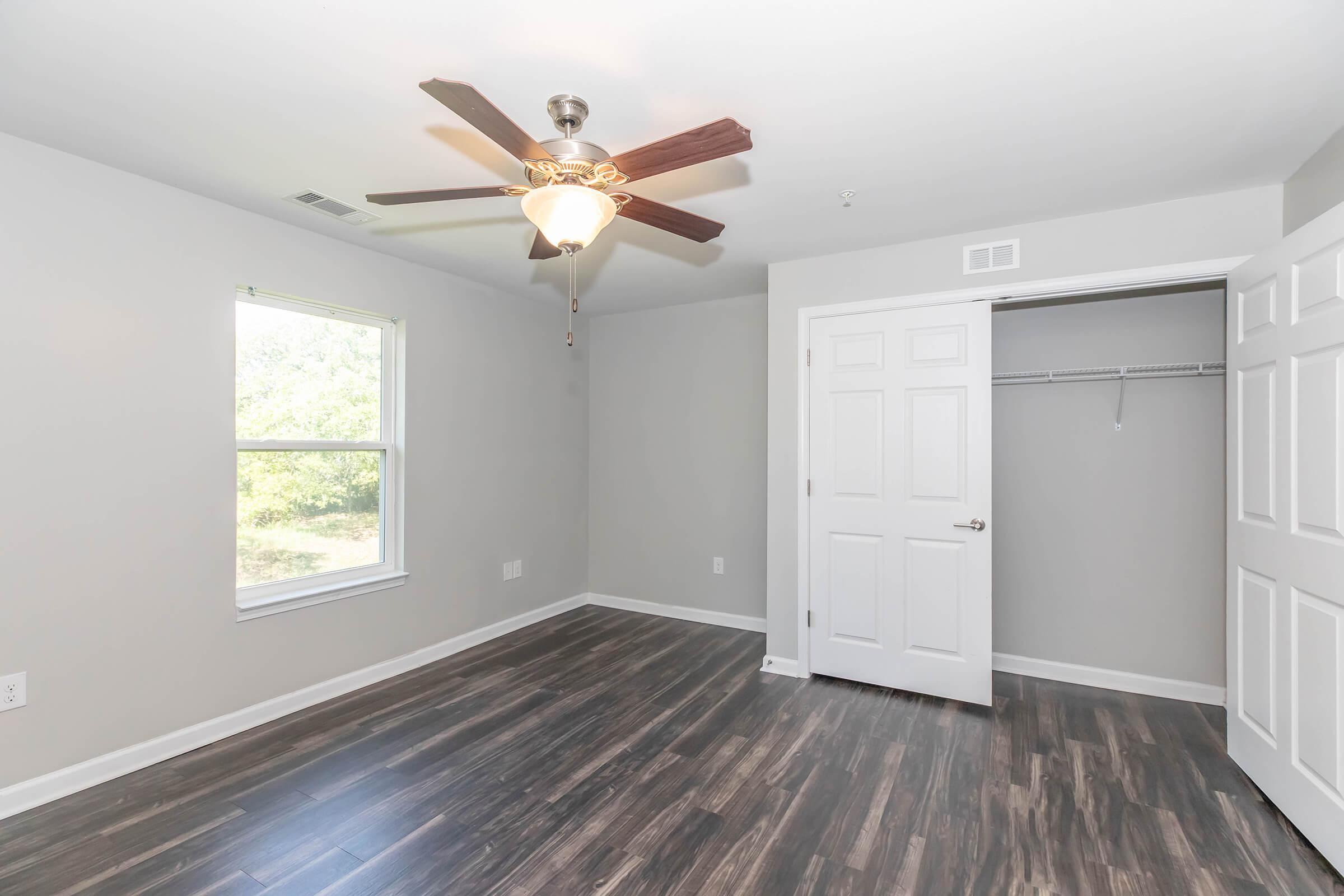 A well-lit, empty room featuring a ceiling fan, large window with a view of greenery, and dark wood-like flooring. A closet with white doors is partially open on the right side, providing additional storage space. The walls are painted in a light gray color, giving the room a spacious and modern feel.