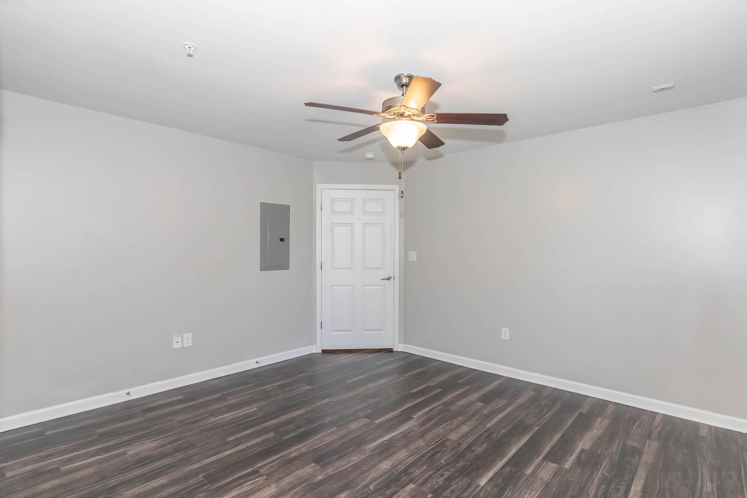 A spacious, well-lit room with a ceiling fan, light gray walls, and dark hardwood-style laminate flooring. The room features a closed white door, an electrical panel on the wall, and a clean, empty layout suitable for various furnishings.
