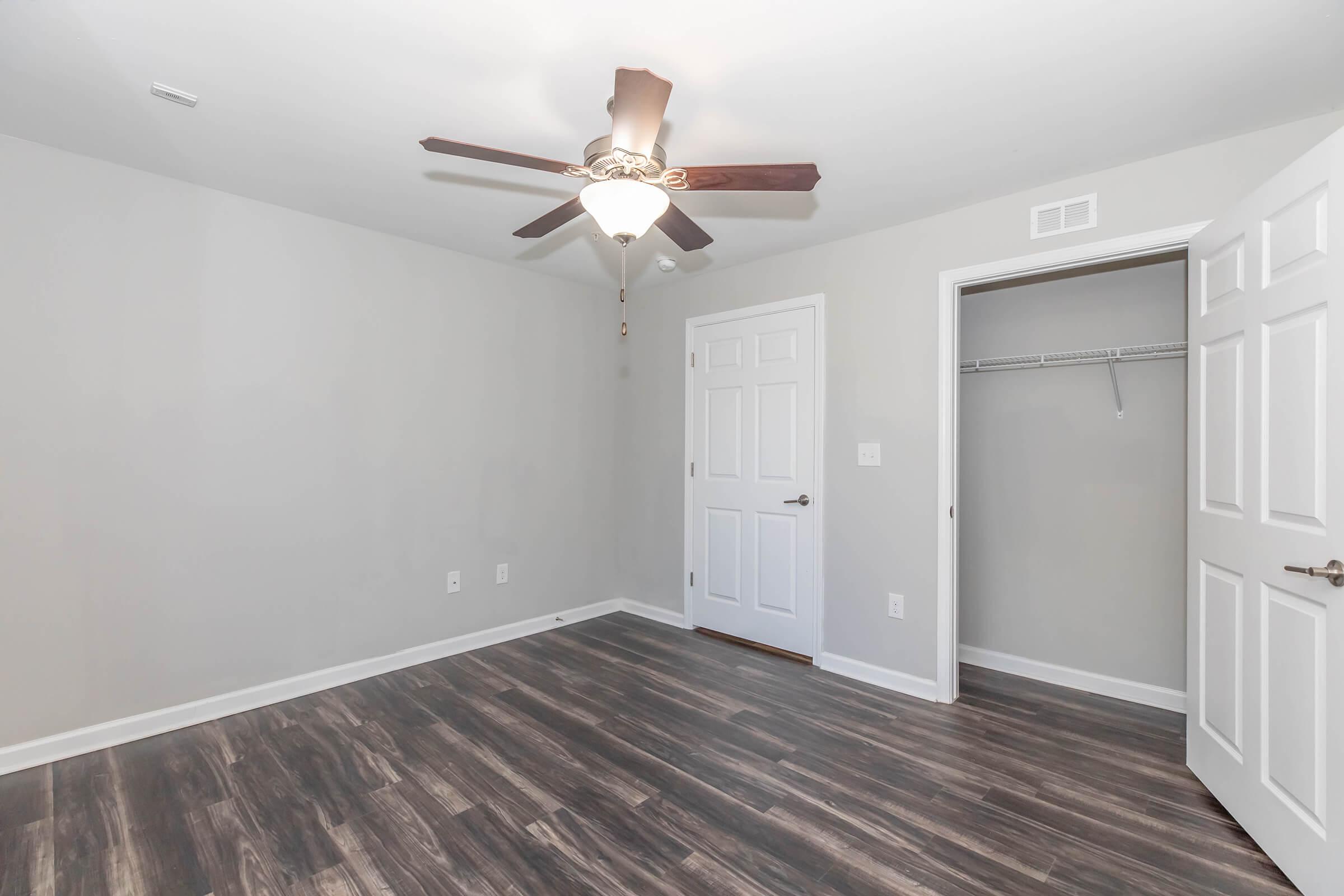 Empty bedroom with light gray walls and dark wood laminate flooring. A ceiling fan with wooden blades is installed in the center. There is a white closet with sliding doors to the right, and a door leading to a hallway on the left. Natural light is coming from the window outside the frame.