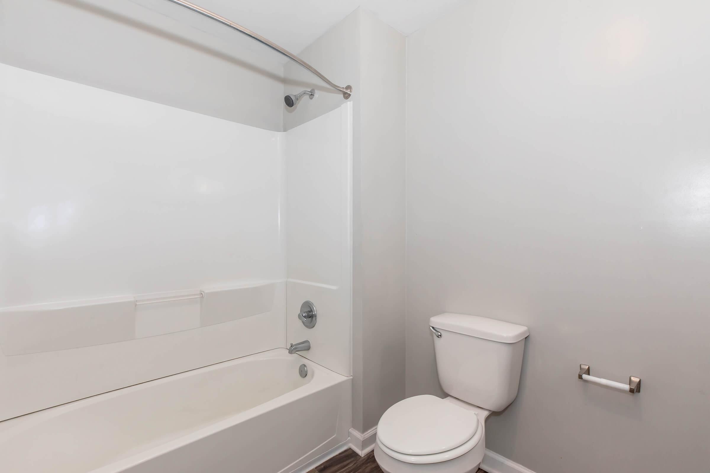A clean, modern bathroom featuring a white bathtub with a shower, a toilet, and light gray walls. The space has a minimalistic design, with a towel holder mounted on the wall and a wooden floor to add a touch of warmth. Natural light enhances the bright and airy atmosphere.