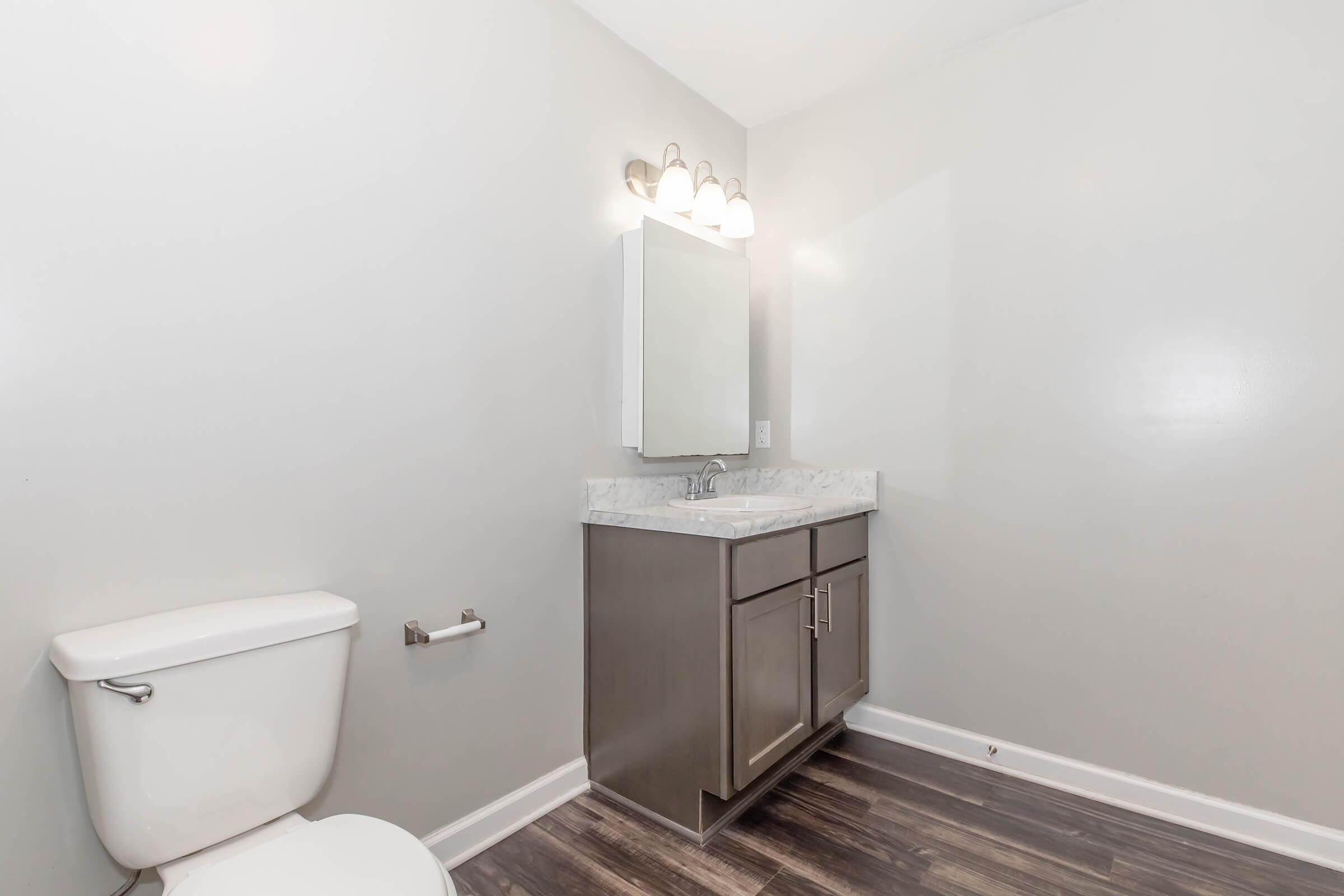 A modern bathroom featuring a white toilet, a sleek vanity with a marble countertop, a wall-mounted mirror, and three light fixtures. The walls are painted a light gray, and the flooring is dark laminate wood. The space is well-lit and clean, creating a contemporary aesthetic.