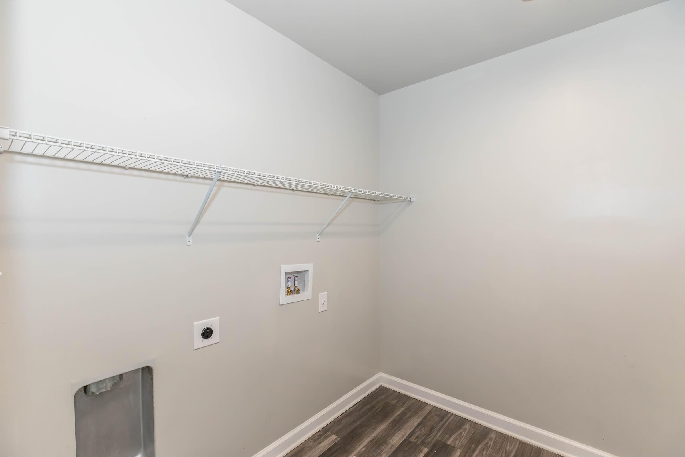 A clean, empty laundry room with a light gray wall. A wire shelf runs along one side, and there are electrical outlets and a dryer vent on the wall. The floor features dark wooden planks, adding a modern touch to the space.