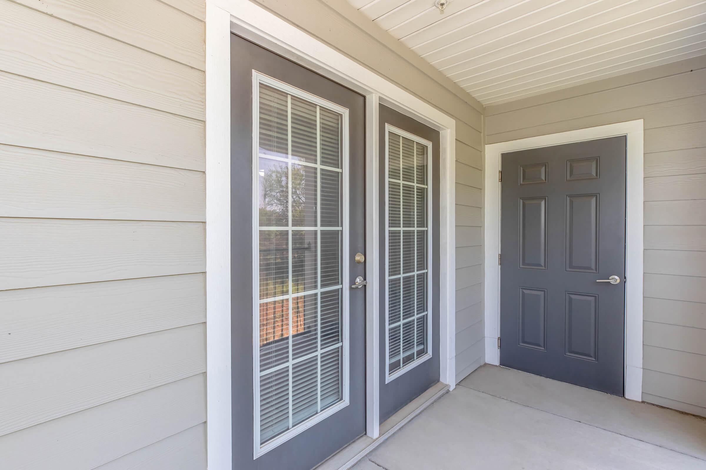 Double glass doors with white frames are set into a light gray wooden exterior wall, complemented by a matching single door nearby. The covered entryway features a concrete floor and is well-lit with natural light.