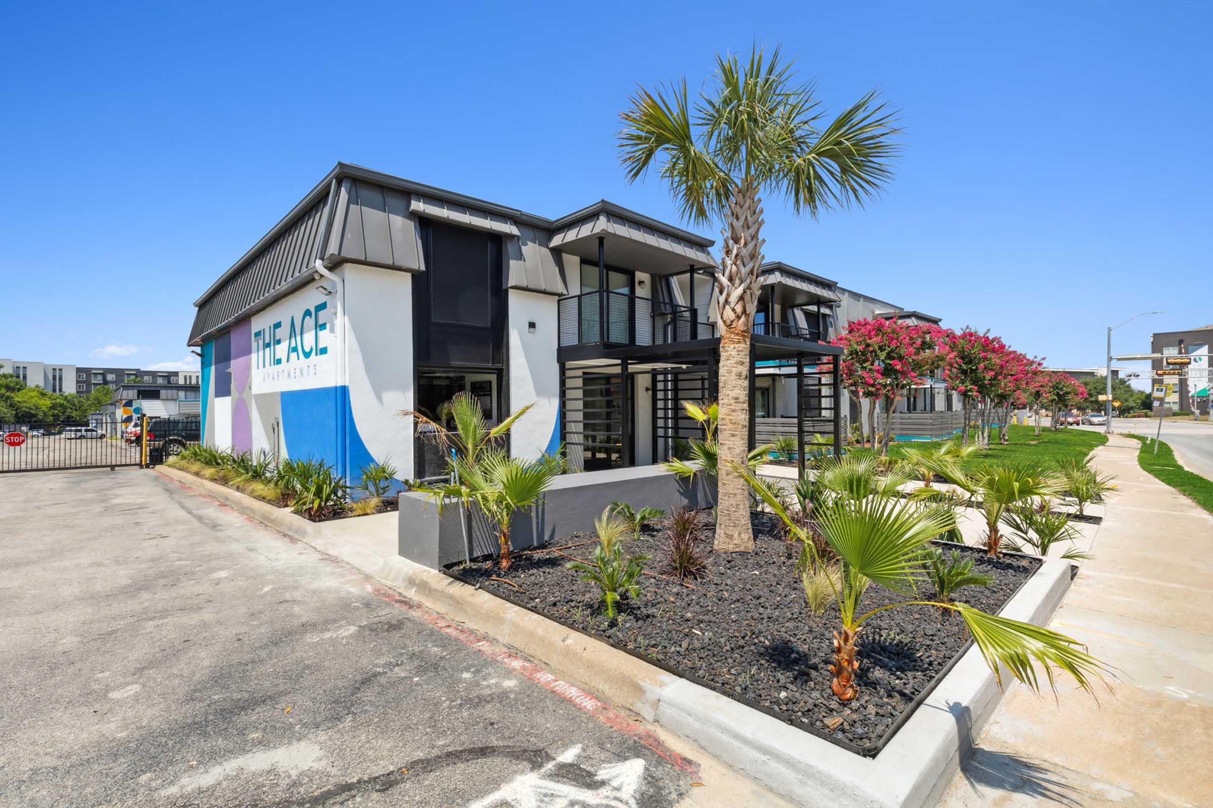 A modern apartment complex named "The Ace Apartments," featuring a stylish exterior with palm trees and colorful murals. The building is well-maintained, with landscaped surroundings and clear blue skies in the background. The entrance is gated, adding an element of security.