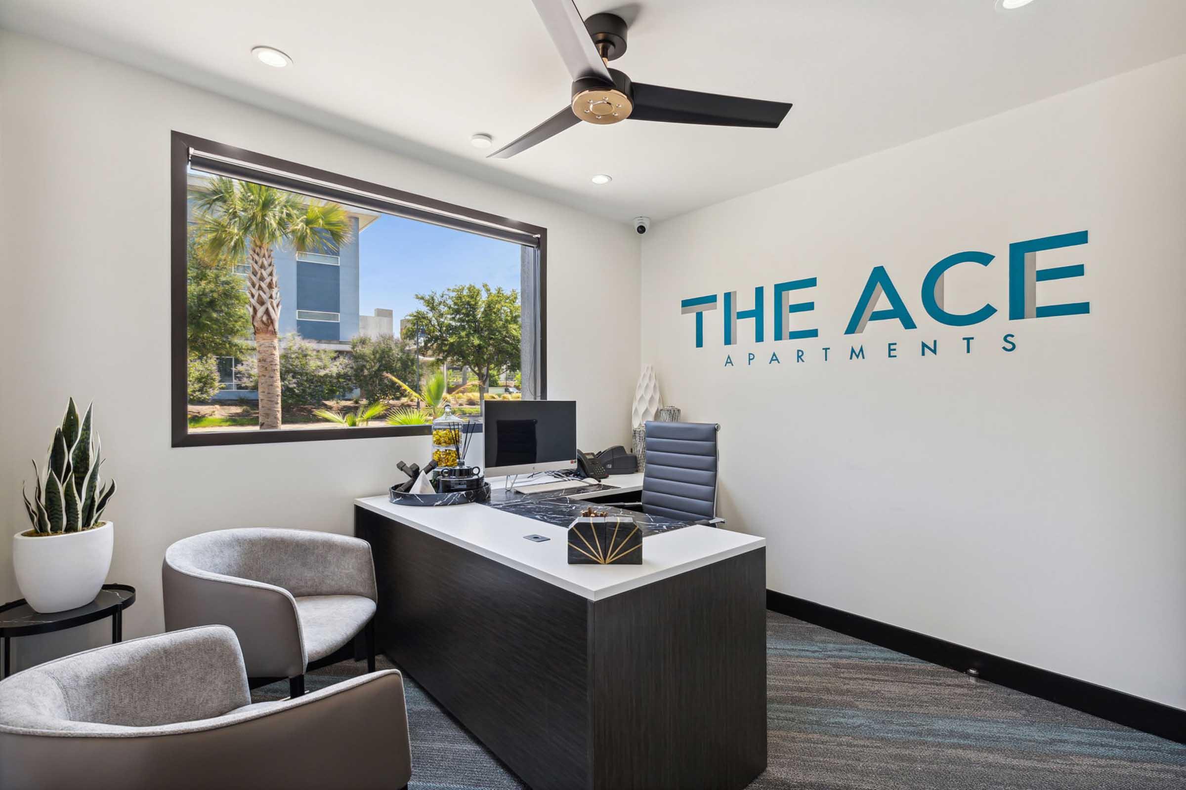 Modern office space with a sleek desk and ergonomic chair. A computer and decorative items are on the desk. To the left, there’s a potted plant, and large windows let in natural light, providing a view of greenery outside. The wall features the text "THE ACE APARTMENTS" in bold, turquoise letters.