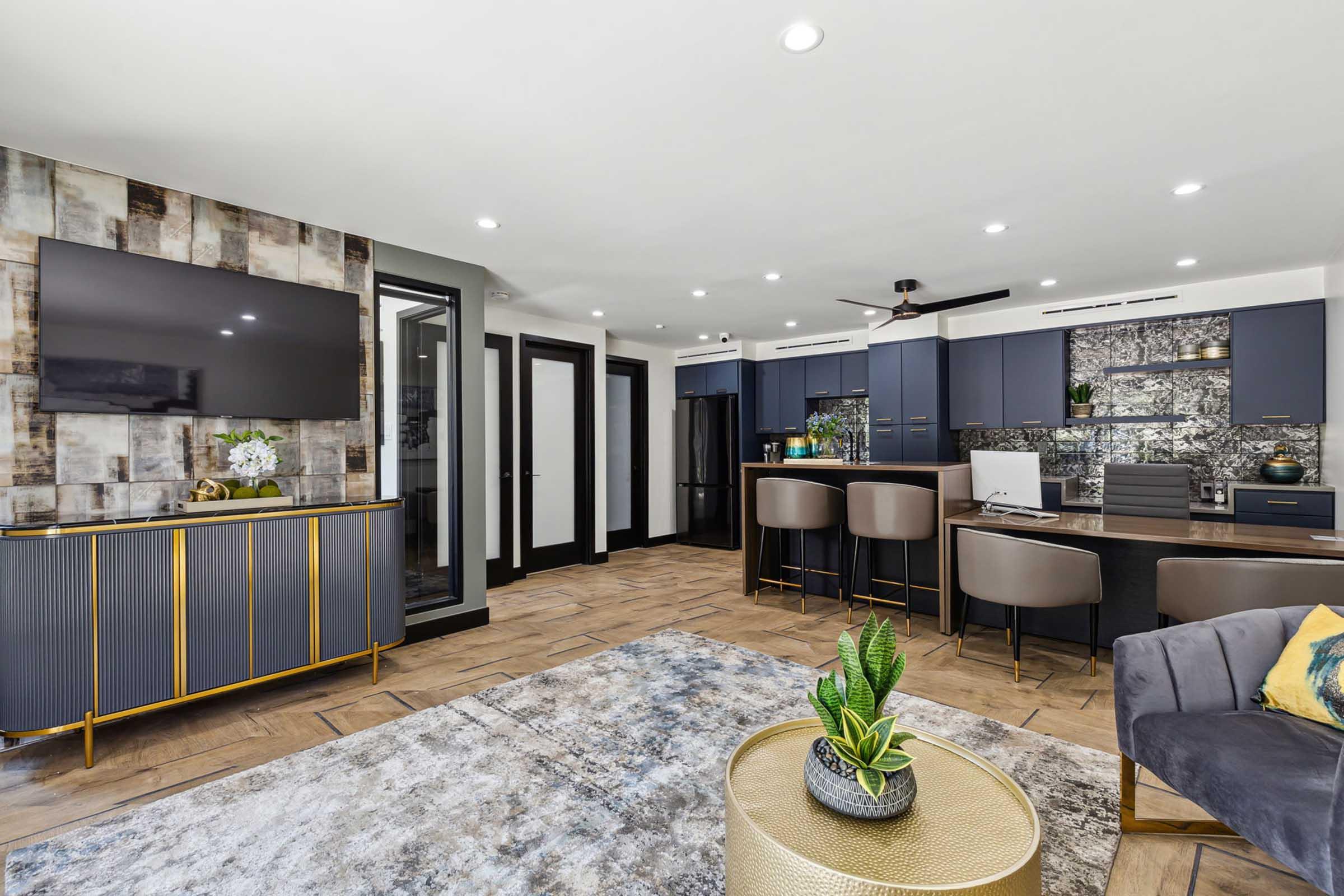 A modern interior featuring an open-plan kitchen and living area. The kitchen has dark cabinetry and a decorative backsplash, alongside a refrigerator. A comfortable seating area includes a gray sofa and a round coffee table with a plant. Soft lighting enhances the contemporary design with a stylish rug underfoot.