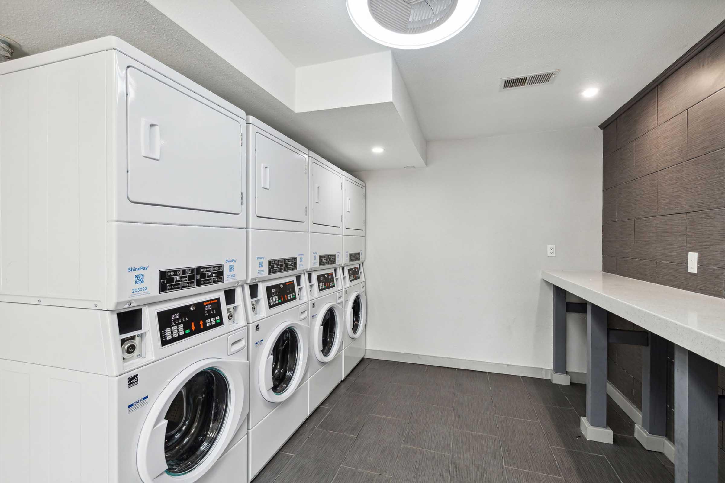 A modern laundry room featuring stacked white washer and dryer units, with a countertop nearby. The walls are painted in light colors and the flooring is dark tiles, creating a clean and contemporary look. Soft overhead lighting enhances the spacious feel of the room.