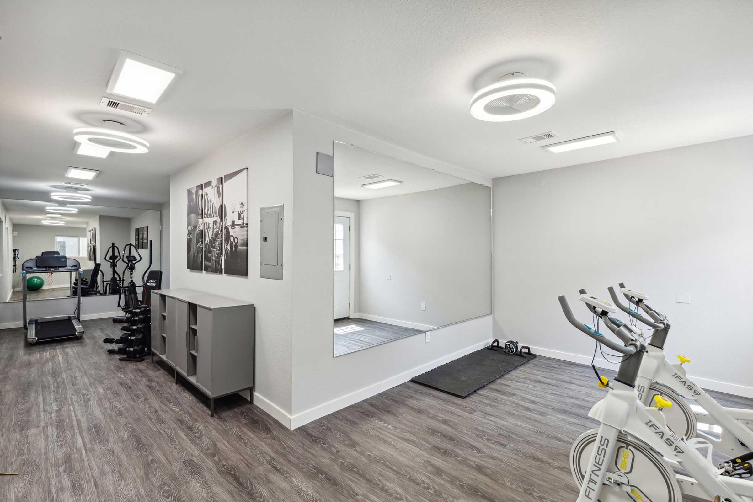 A modern fitness room featuring exercise equipment such as stationary bikes and free weights. Mirrors line one wall to enhance the space, and there are large windows allowing natural light. The flooring is a light wood, and the walls are painted in neutral tones, creating a bright and inviting atmosphere for workouts.
