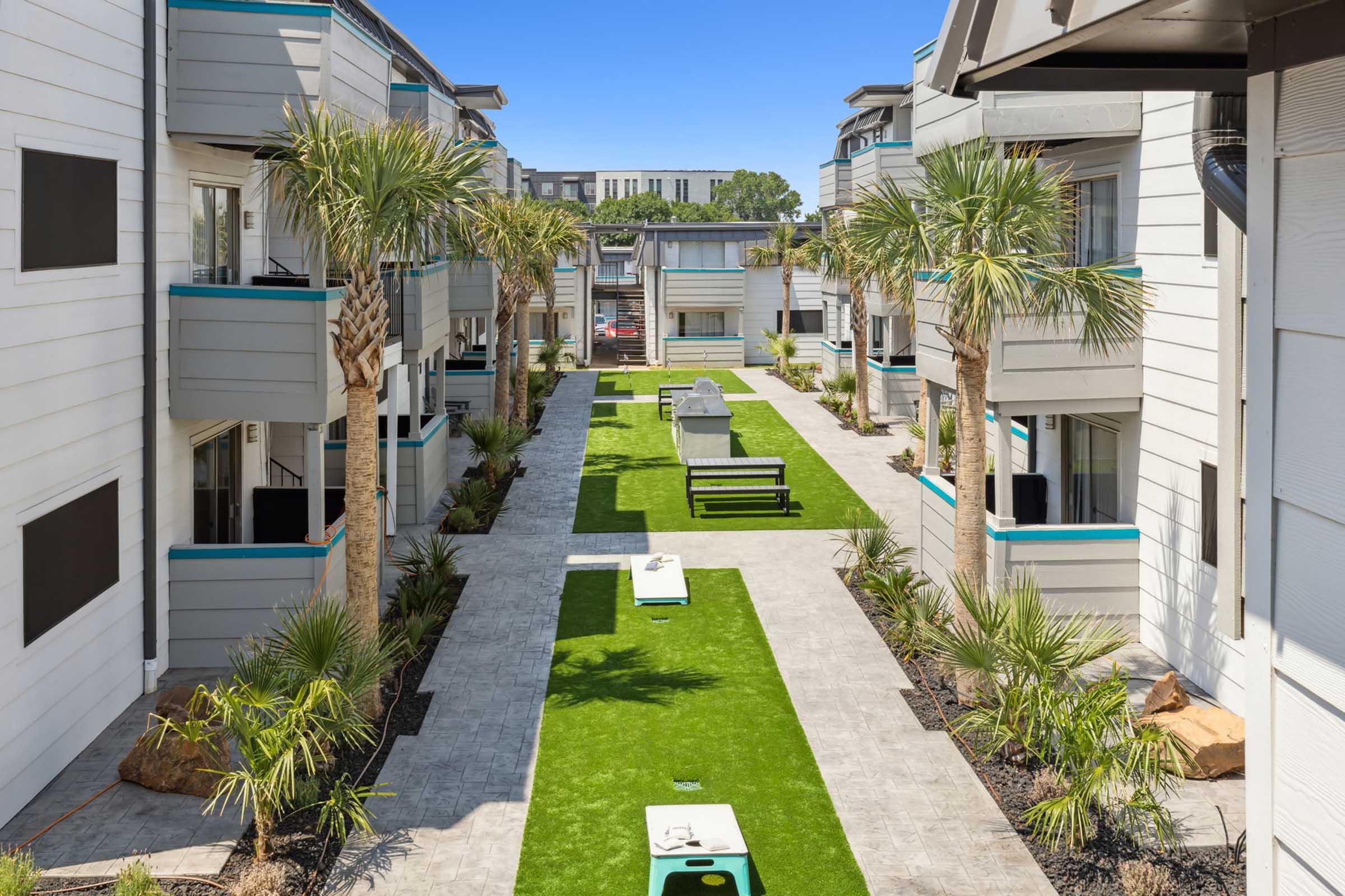 A landscaped courtyard surrounded by modern apartment buildings, featuring palm trees, green grass, and benches. The pathway is paved with stone, and there are amenities like a barbecue grill and cornhole game setup in the outdoor space. Clear blue skies complete the scene.