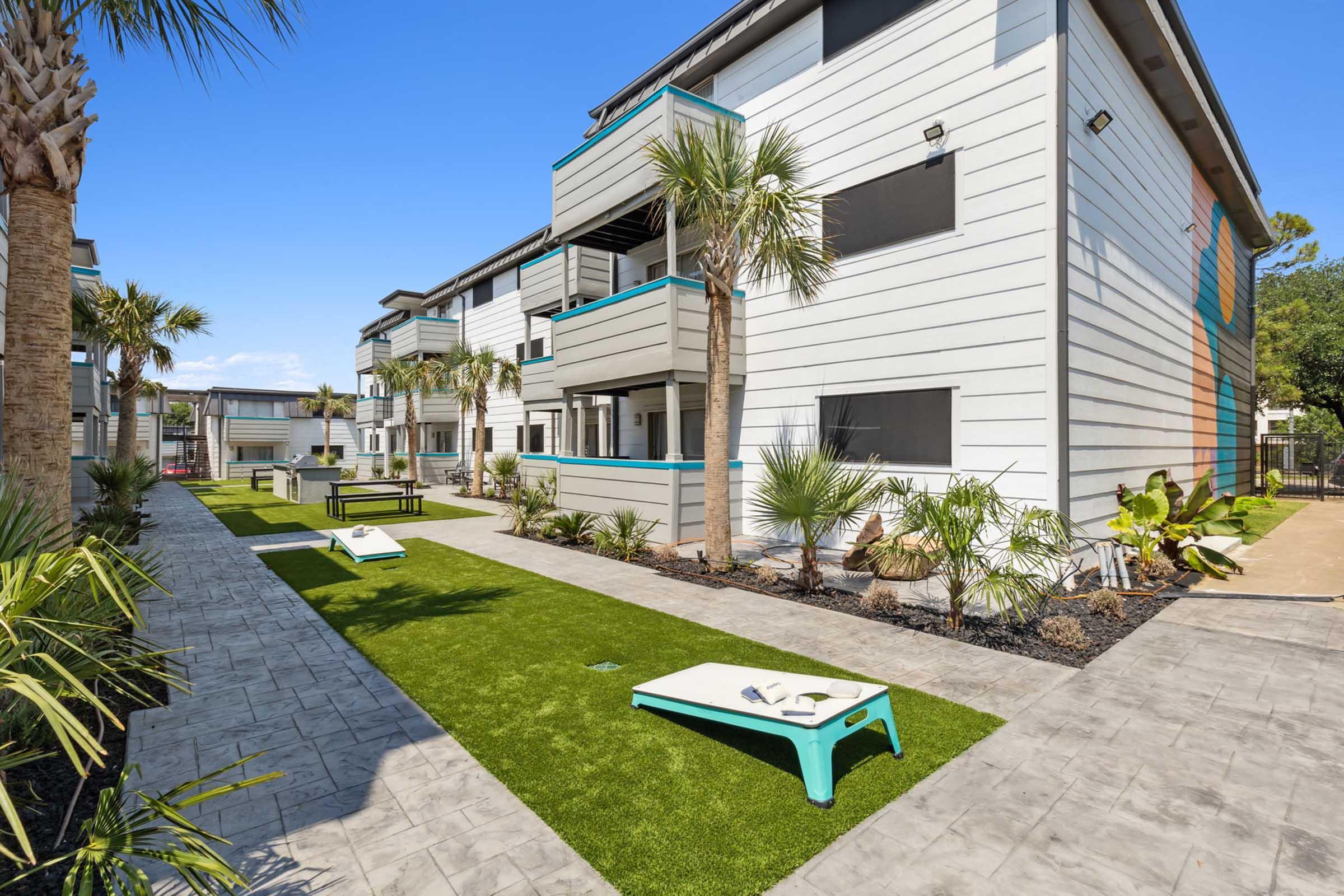 A modern apartment complex featuring two buildings with palm trees and well-maintained landscaping. The grounds include pathways of artificial grass, decorative plants, and areas for outdoor games. Bright blue accents on the balconies provide a lively contrast against the white and gray exterior walls.