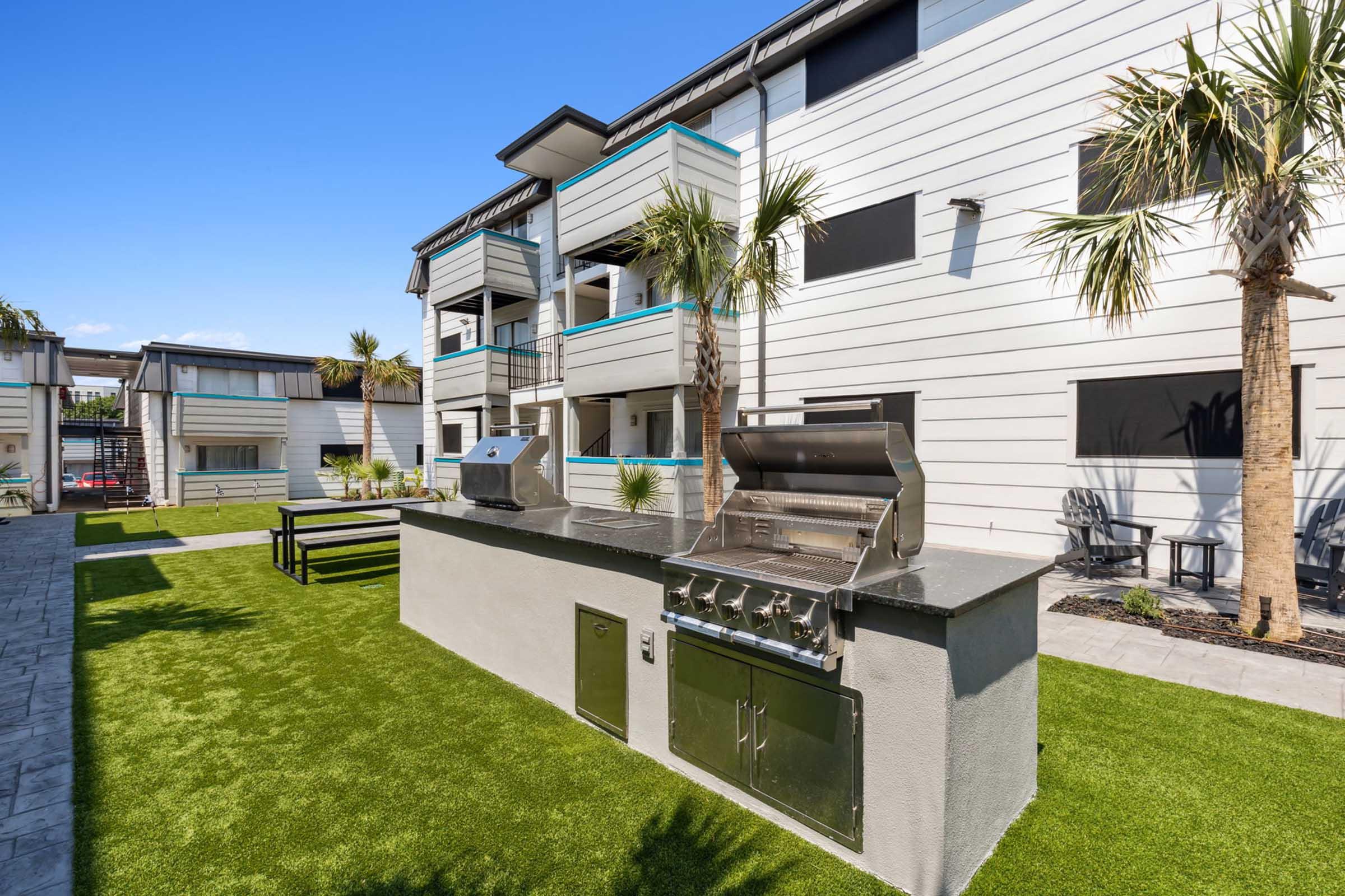 An outdoor grilling area featuring a stainless steel barbecue grill on a concrete island, surrounded by green artificial grass. In the background, there are modern apartment buildings with balconies and palm trees, under a clear blue sky.