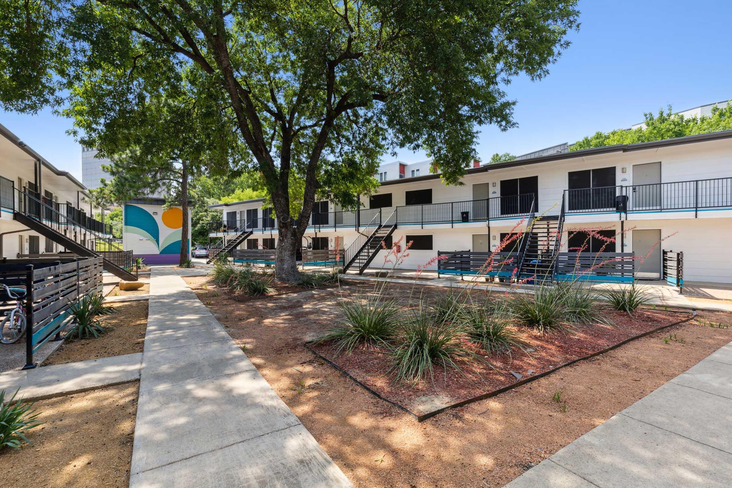 A colorful courtyard of a two-story apartment complex featuring trees, landscaped gardens, and a mural on one building. Walkways connect the entrances, with stairs leading to the second floor, and bicycles parked in the foreground. The scene is set under a clear blue sky.