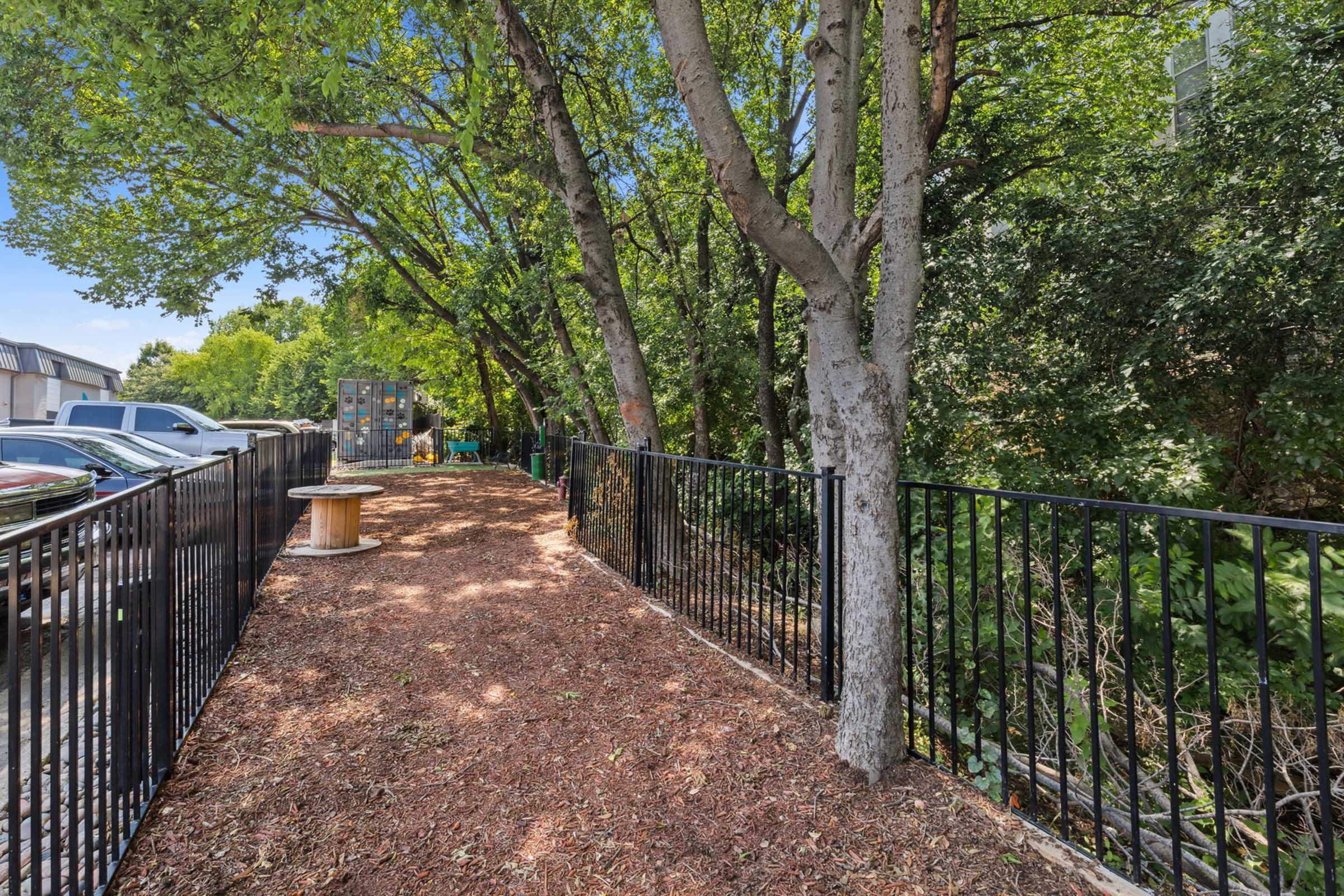A pathway surrounded by trees and lush greenery, featuring a wooden spool and a black metal fence. The area has a natural, rustic feel, with mulch covering the ground. Cars are parked nearby, and there's a glimpse of a storage or utility shed in the background.
