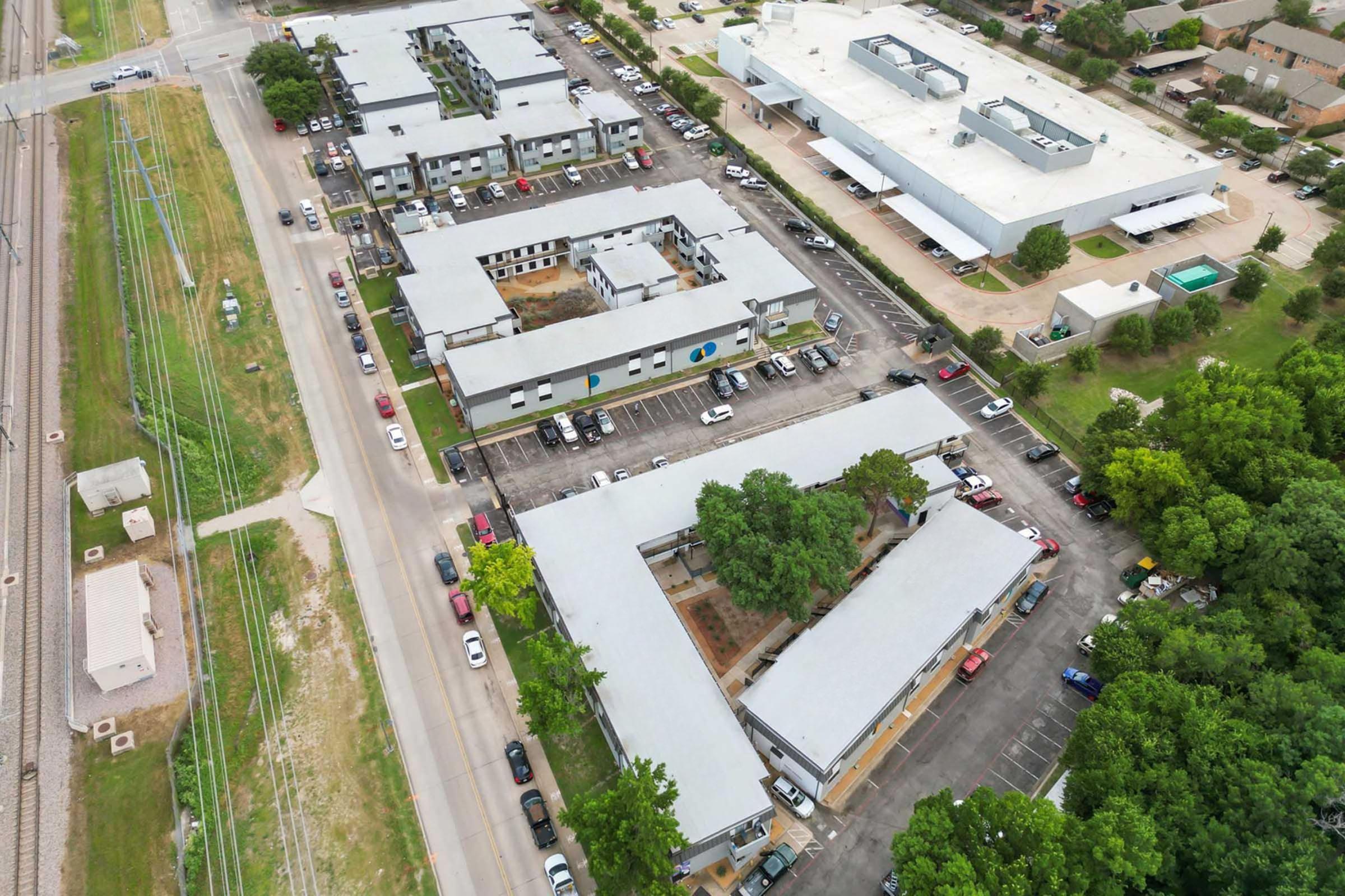 Aerial view of a residential complex featuring multiple low-rise buildings and parking lots. Surrounding greenery and trees, with a nearby road and railway visible. The scene captures a mix of urban and natural elements, showcasing the layout of the properties and nearby amenities.