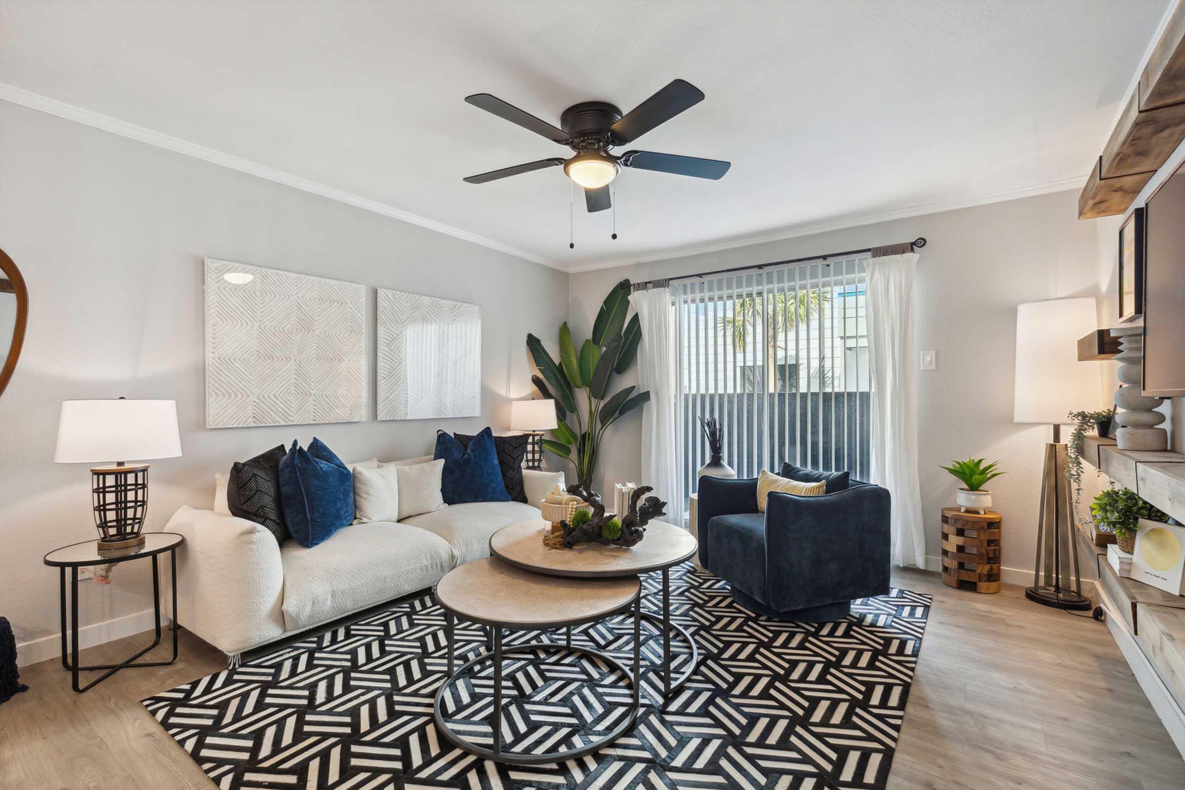 Cozy living room featuring a white sofa with blue pillows, a round coffee table, and a patterned black and white area rug. Two lamps illuminate the space, complemented by plants and decorative accents. A ceiling fan and large window provide light and ventilation, creating a welcoming atmosphere.