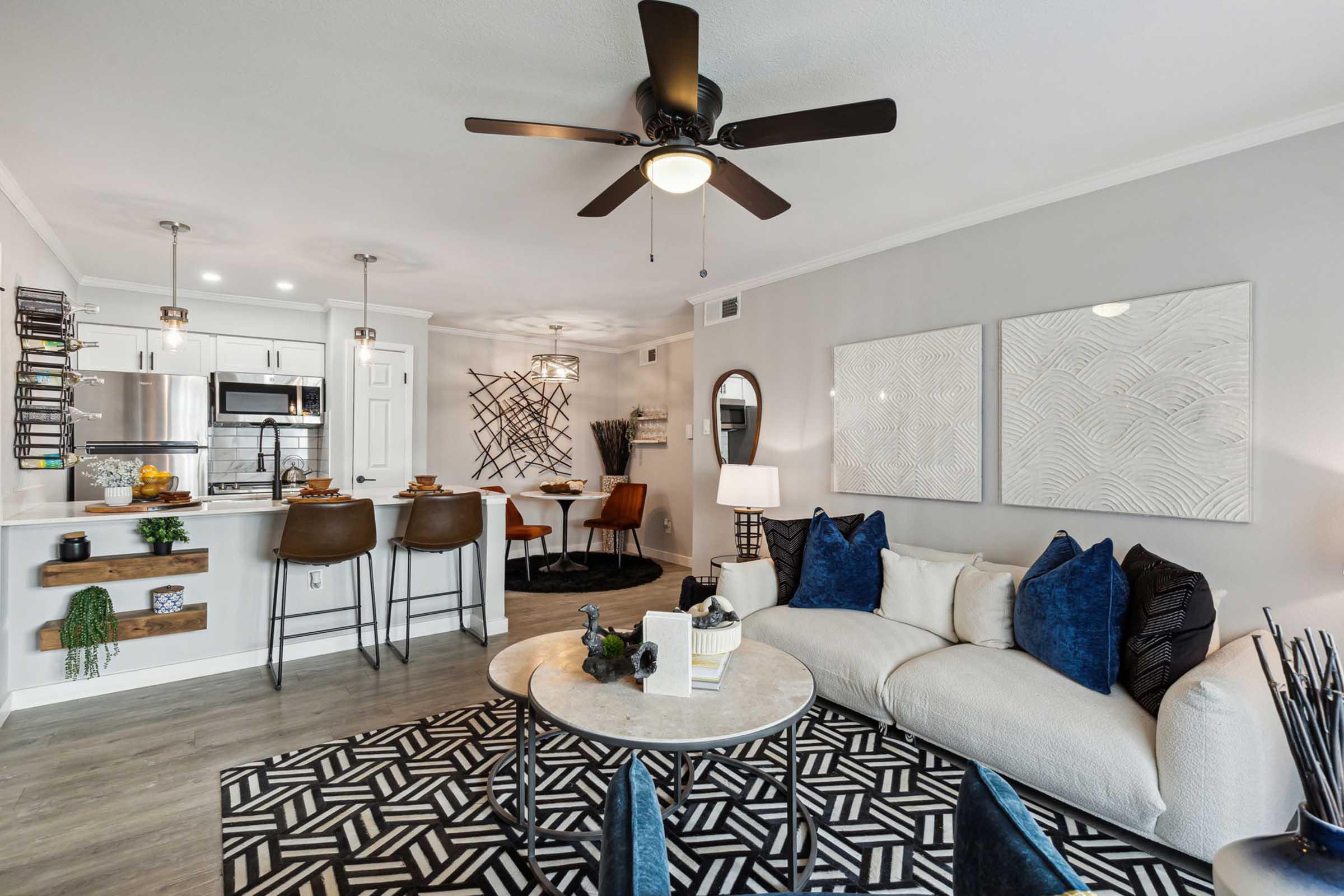 A modern living room and kitchen area featuring a cozy white sofa with blue cushions, a round table with brown bar stools, and stylish decor. The space has a ceiling fan and textured artwork on the walls, along with a geometric black and white area rug enhancing the contemporary aesthetic.