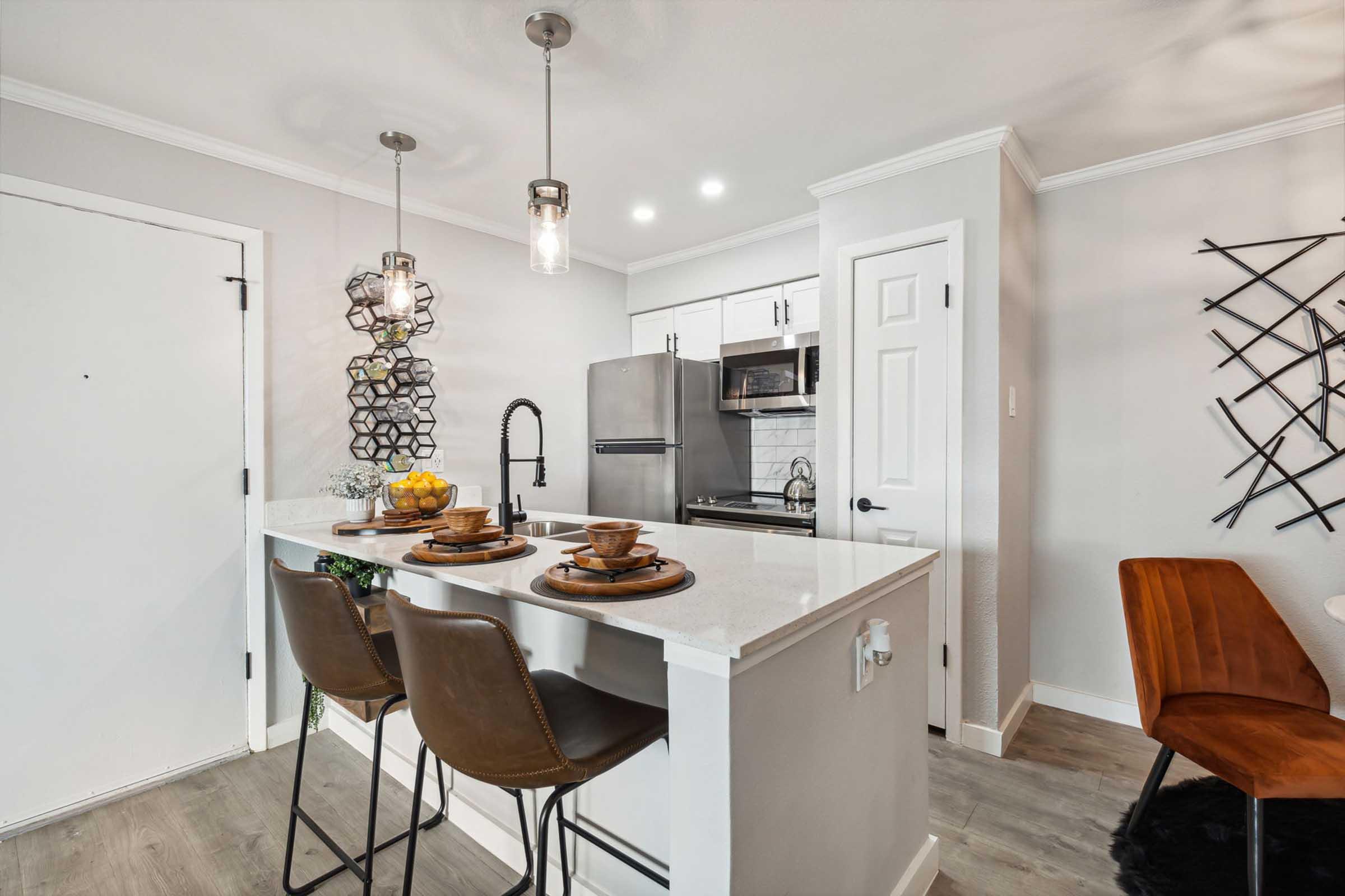 A modern kitchen featuring a white island with two brown bar stools, elegant pendant lighting, and a sleek black faucet. The area has stylish decor, including a fruit bowl and decorative wall pieces, with stainless steel appliances visible in the background. The walls are painted in soft gray tones.