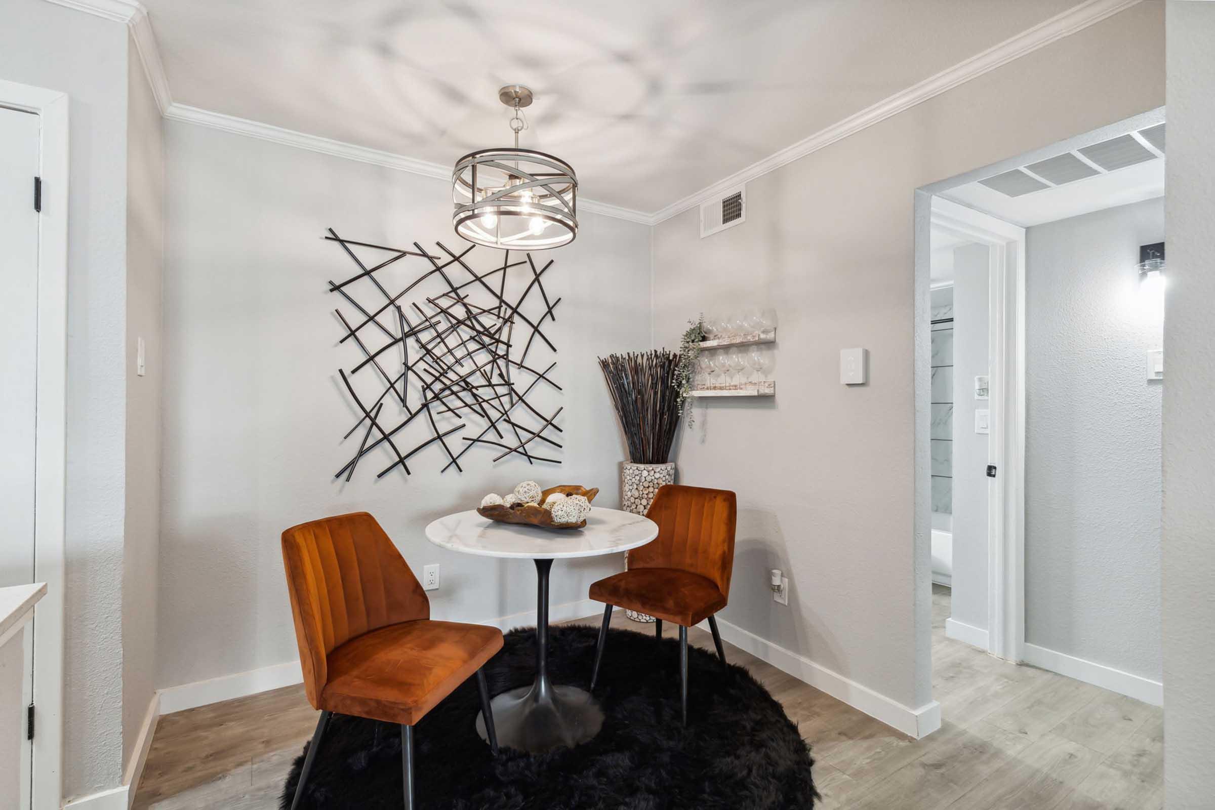 A modern dining area features a round white table with two rust-colored chairs on a black circular rug. A stylish chandelier hangs above, and a decorative wall piece made of intersecting lines is displayed. Decorative elements, including a tall vase with branches, are present in the corner.