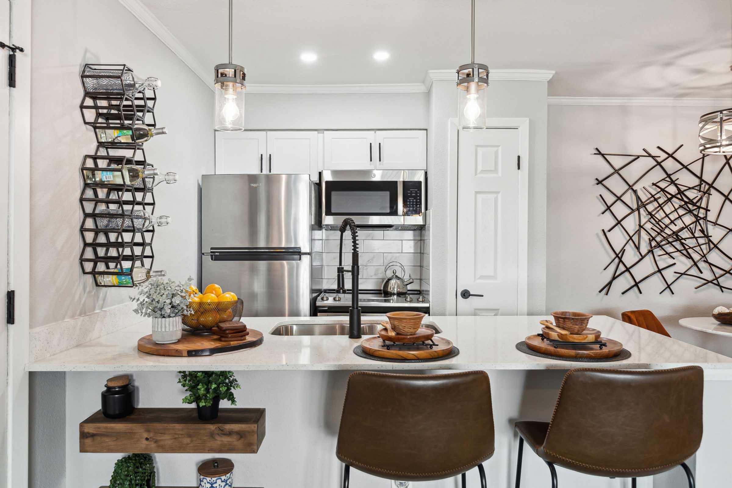 A modern kitchen featuring a countertop with brown bar stools, plates, and utensils. The kitchen has stainless steel appliances, including a refrigerator and microwave, and is accented with decorative items like a plant and a wire shelf holding small containers. Natural light illuminates the space.