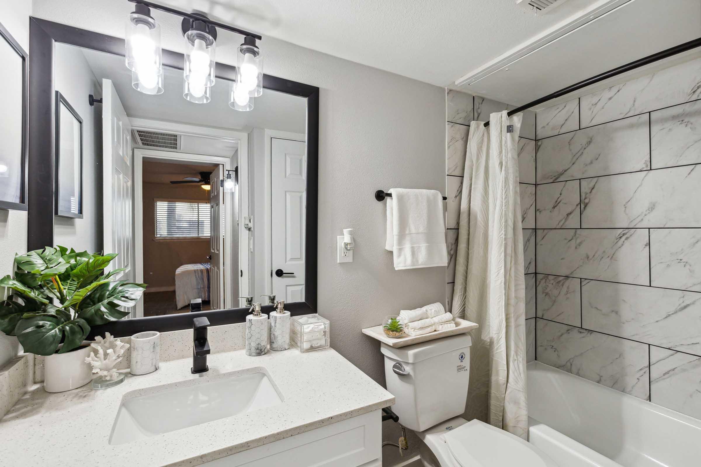 Modern bathroom featuring a white countertop sink with a black faucet, a large mirror with four light fixtures above, and a bathtub with a shower curtain. Decor includes a potted plant and neatly arranged towels. The walls are tiled with a marble design, creating a clean and stylish atmosphere.