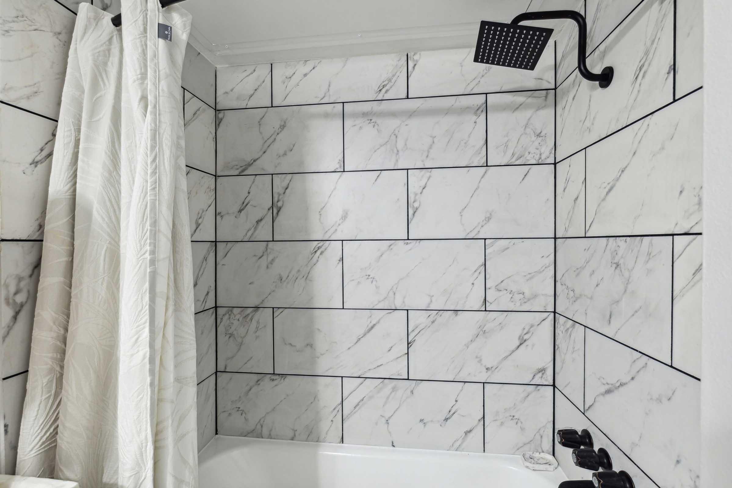 A tiled bathroom shower area featuring modern marble-style tiles. There is a sleek black showerhead mounted on the wall and a white shower curtain partially visible, adding a clean and contemporary aesthetic to the space.