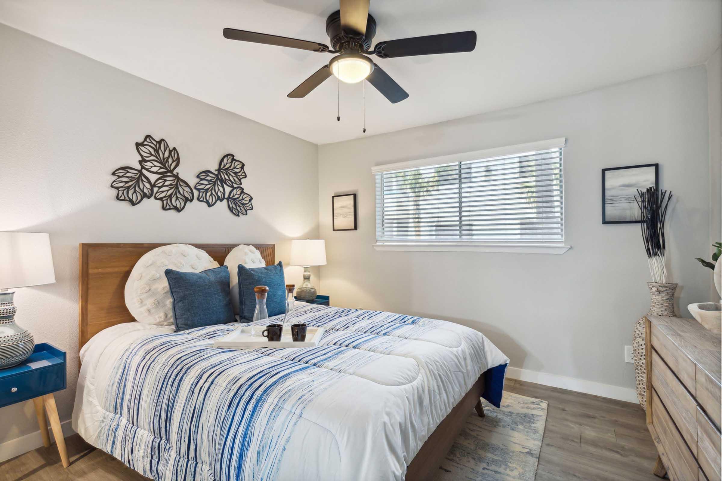 A cozy bedroom featuring a queen-sized bed with a blue and white striped comforter, two bedside lamps, a dresser, and decorative wall art. A ceiling fan is overhead, and there is a window with blinds allowing natural light to brighten the room.