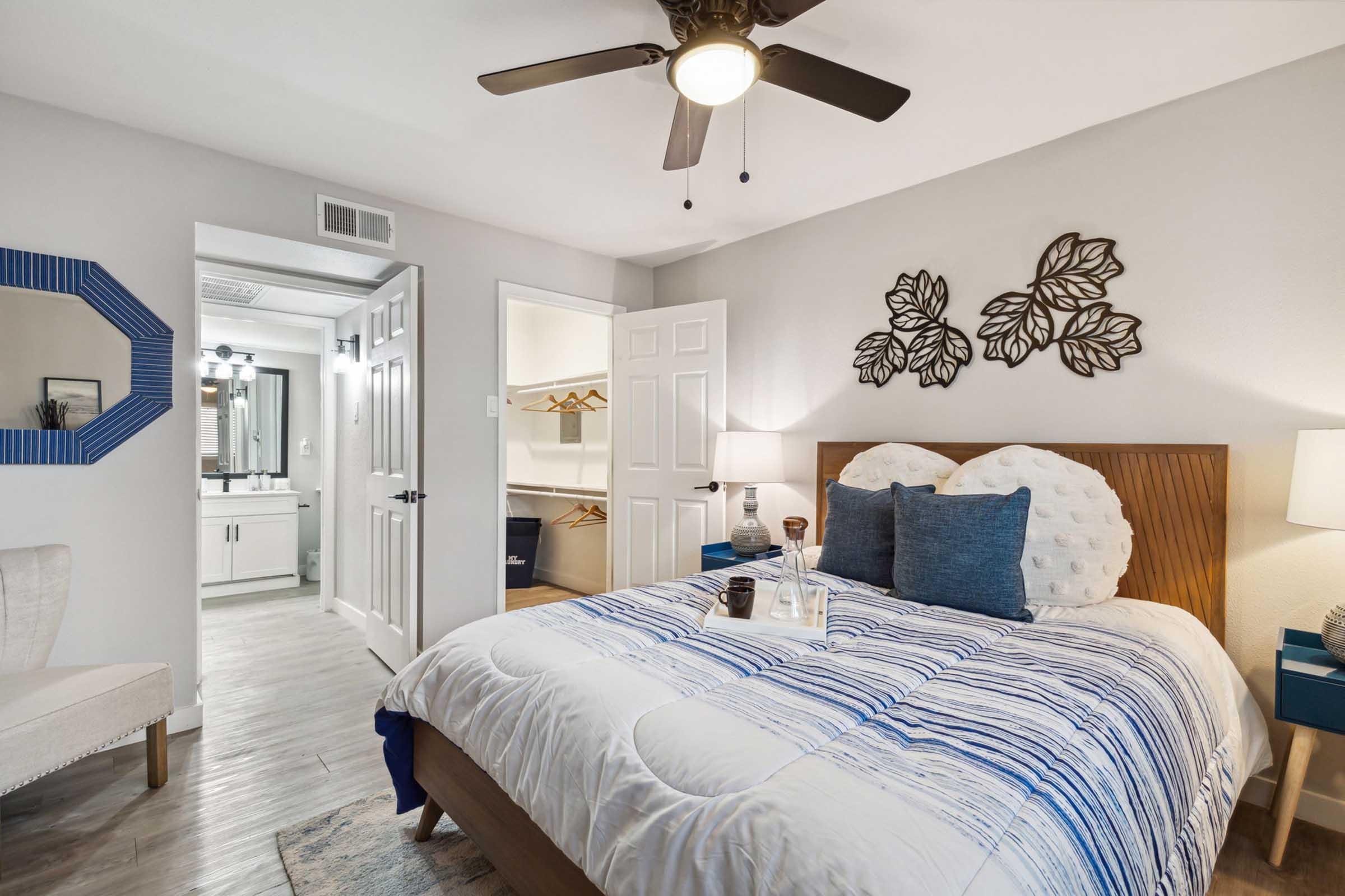 A cozy bedroom featuring a bed with a blue and white striped comforter, two decorative pillows, and a wooden headboard. There is a ceiling fan, side tables with lamps, and a chair in the corner. A closet and a bathroom are visible through two open doors. The room has modern decor with leaf wall art.