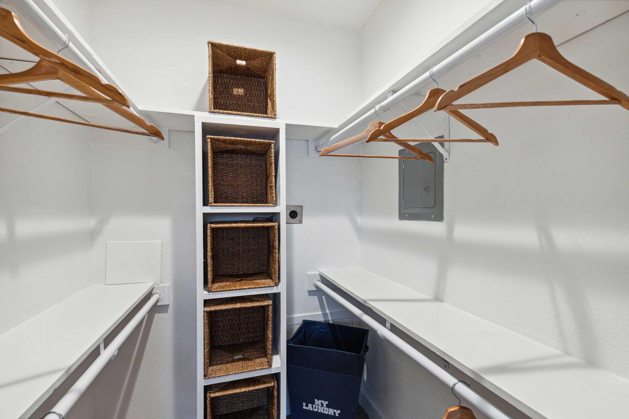 A well-organized closet with white walls and wooden hangers. There are empty shelves on both sides, stacked wicker baskets in the center, and a blue laundry basket at the bottom. The space is bright and tidy, ideal for storing clothes and accessories.