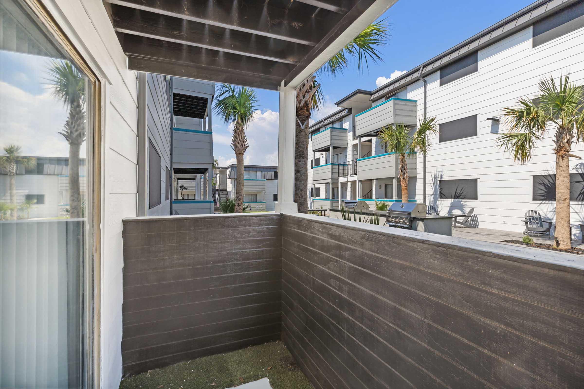 A view from a balcony showcasing a wooden railing, with palm trees in the background. The scene includes modern apartment buildings under a clear blue sky, with a pathway visible in front. The balcony is partially enclosed, creating a cozy outdoor space.