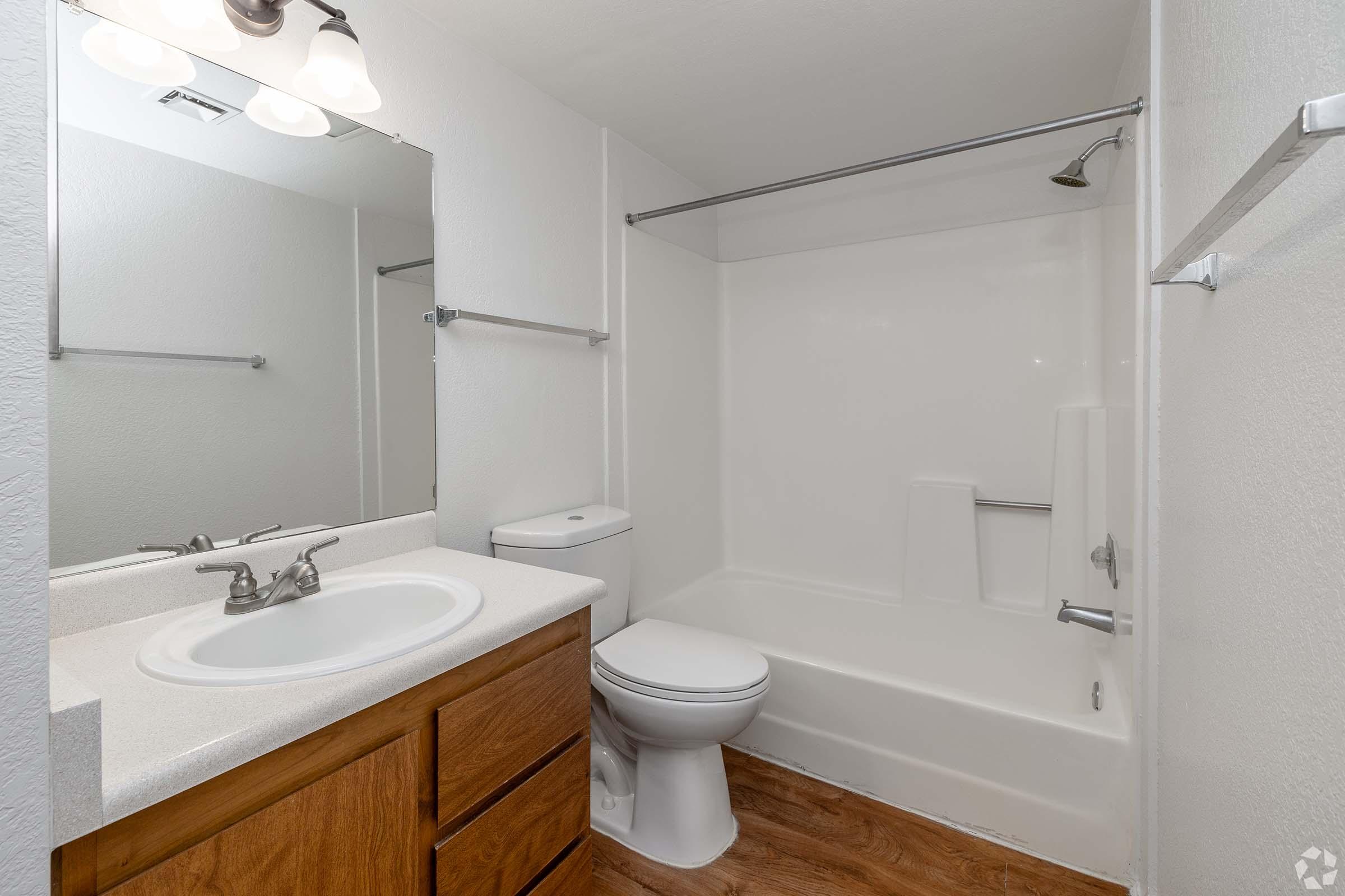 A clean and modern bathroom featuring a bathtub with shower, a toilet, and a single sink vanity with a mirror above. The walls are painted white, and there is wooden flooring. The overall appearance is bright and well-maintained.